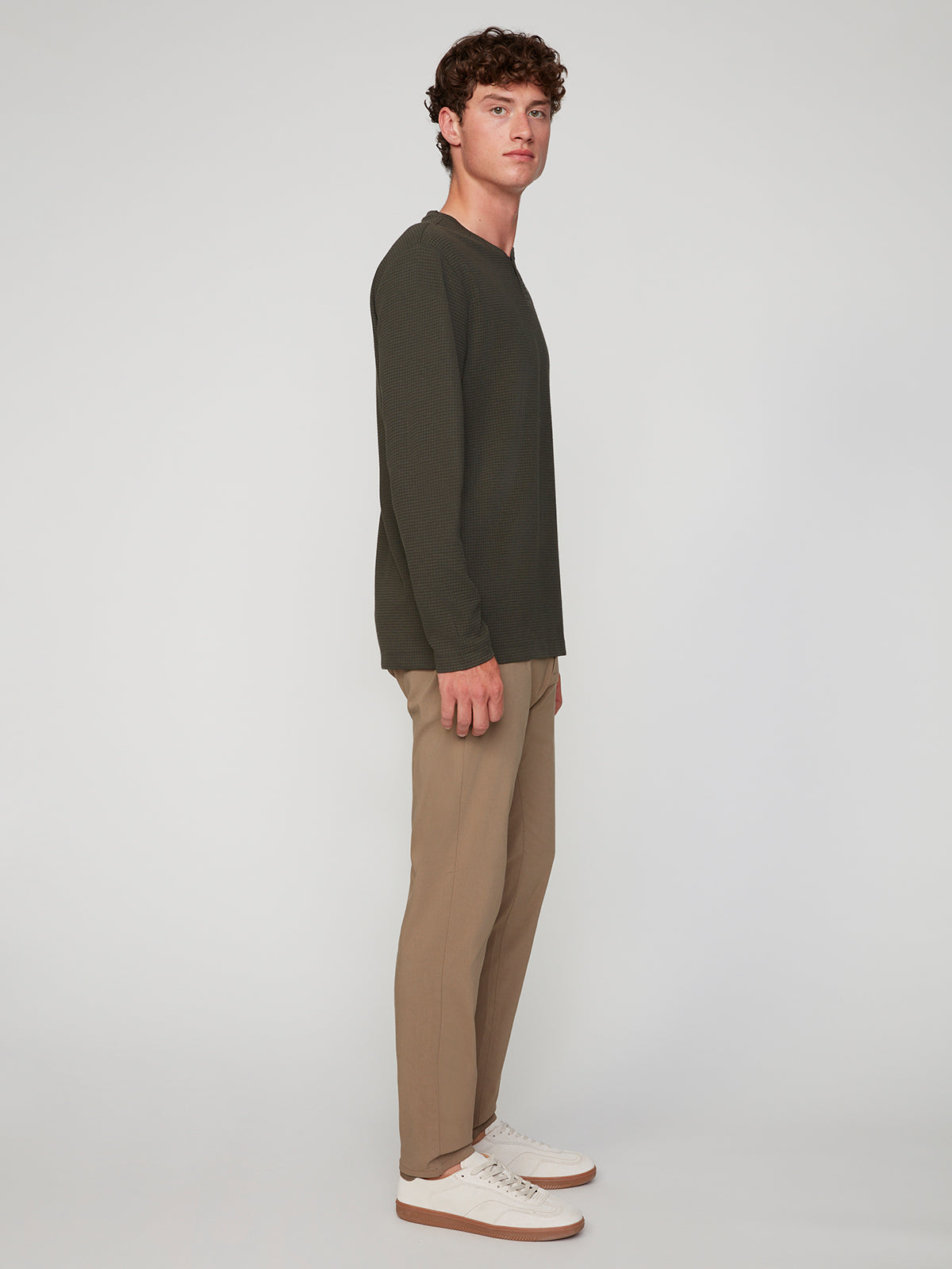 A young man stands in profile against a plain background, wearing a dark long-sleeve shirt, PROJEK RAW MENS 5-POCKET SOLID BUNGEE CHINO PANT, and white sneakers.
