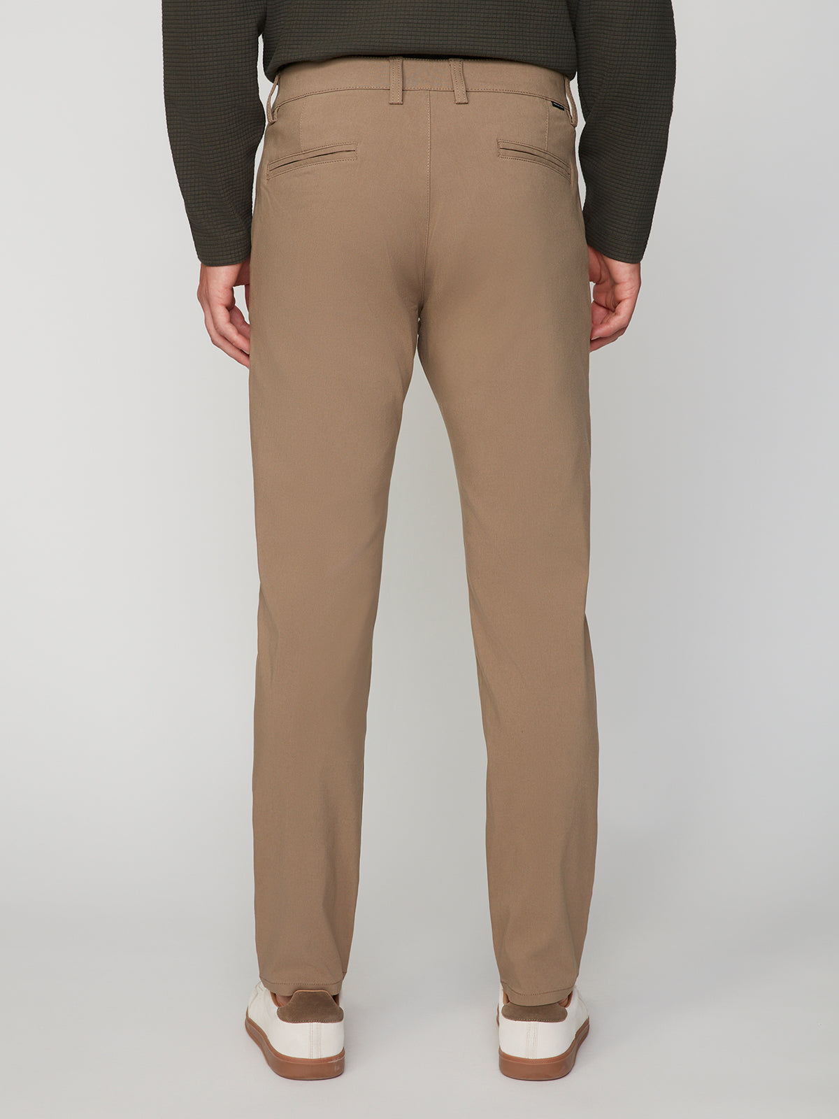 A person is shown standing and facing away from the camera against a plain white background, wearing PROJEK RAW MENS 5-POCKET SOLID BUNGEE CHINO PANT in light brown, paired with a dark long-sleeved shirt and white sneakers.
