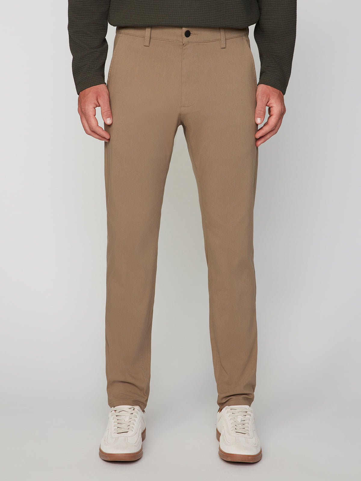 A person wears PROJEK RAW MENS 5-POCKET SOLID BUNGEE CHINO PANT in tan, paired with a dark long-sleeve shirt and white sneakers. The image is cropped at mid-torso on a plain light background, with the face not shown.