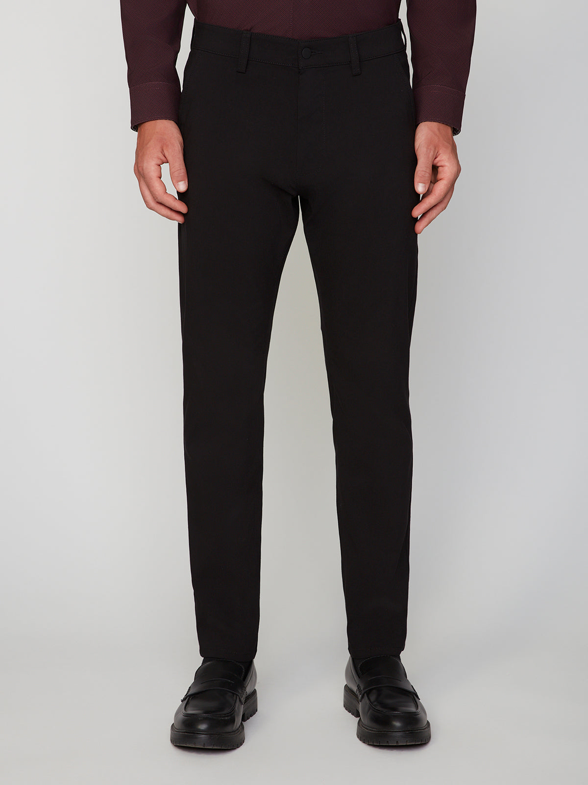 A person is shown from the shoulders down wearing PROJEK RAW MENS 5-POCKET SOLID BUNGEE CHINO PANT in black, paired with a dark long-sleeve shirt and black dress shoes, set against a plain light background.
