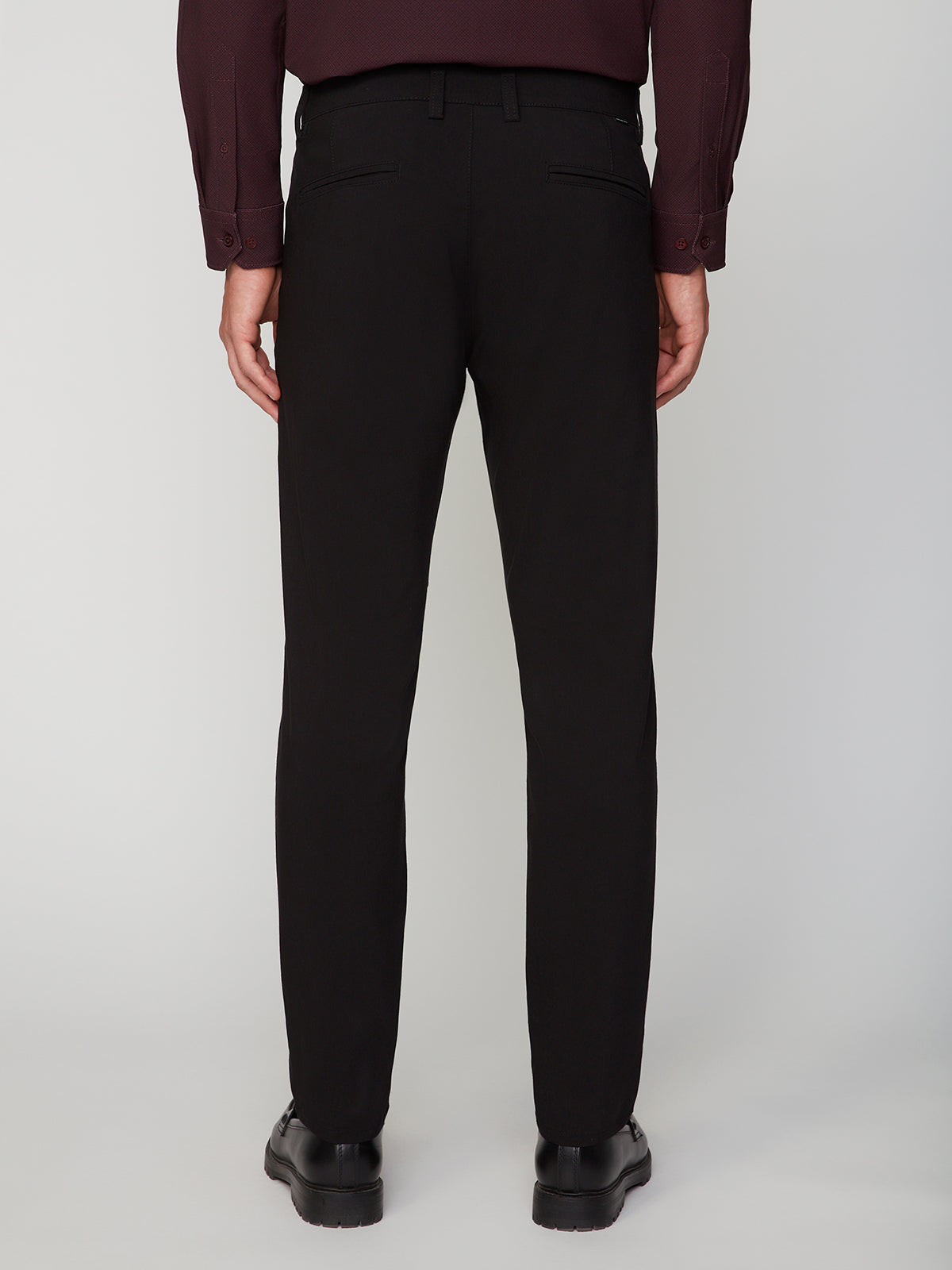 A person wearing PROJEK RAW MENS 5-POCKET SOLID BUNGEE CHINO PANT, black shoes, and a dark long-sleeve shirt stands facing away from the camera against a plain light background.