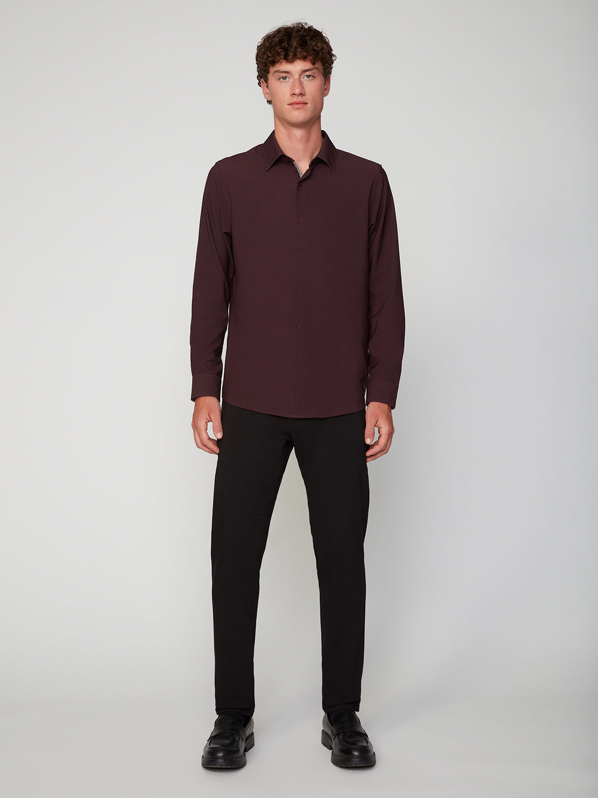 A man stands facing forward against a plain light background, wearing a dark burgundy long-sleeve button-up shirt, PROJEK RAW MENS 5-POCKET SOLID BUNGEE CHINO PANT in black, and matching black shoes.
