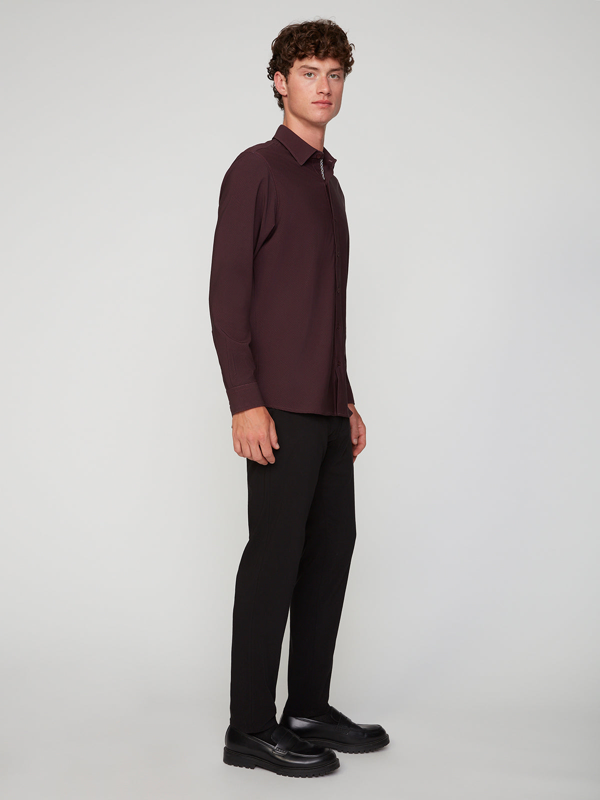 A man with curly brown hair stands in a studio, wearing a long-sleeve dark maroon shirt, PROJEK RAW MENS 5-POCKET SOLID BUNGEE CHINO PANT, and black shoes, facing slightly to the side against a plain light background.