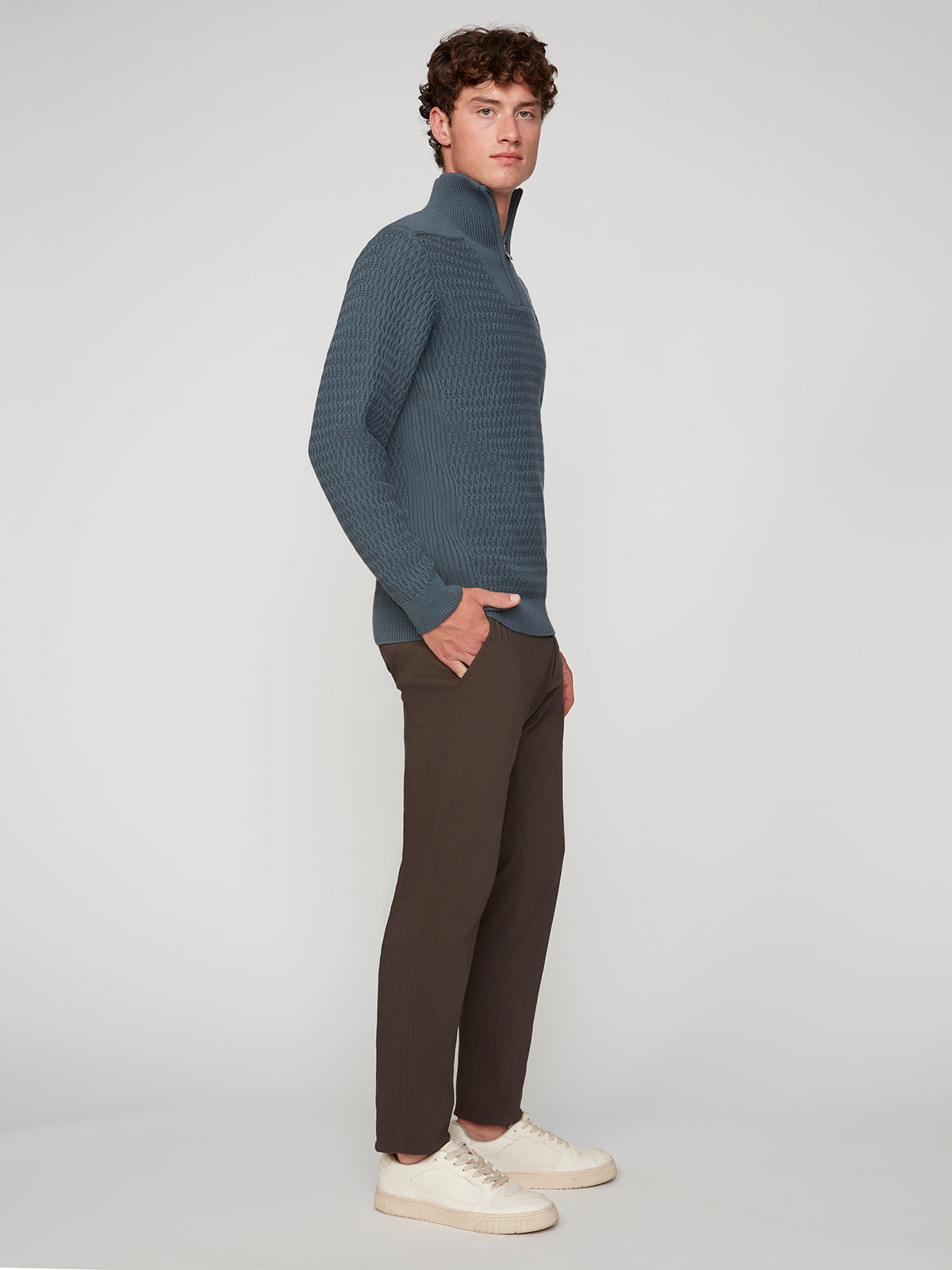 A young man stands sideways, hands in pockets, wearing a textured blue pullover, PROJEK RAW MENS 5-POCKET SOLID BUNGEE CHINO PANT, and white sneakers against a plain light gray background.