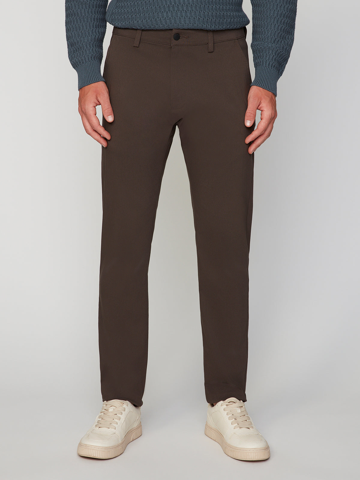 A person wearing PROJEK RAW MENS 5-POCKET SOLID BUNGEE CHINO PANT in a classic fit, with a blue textured sweater and white sneakers, stands against a light gray background. Only the body from shoulders down is shown.