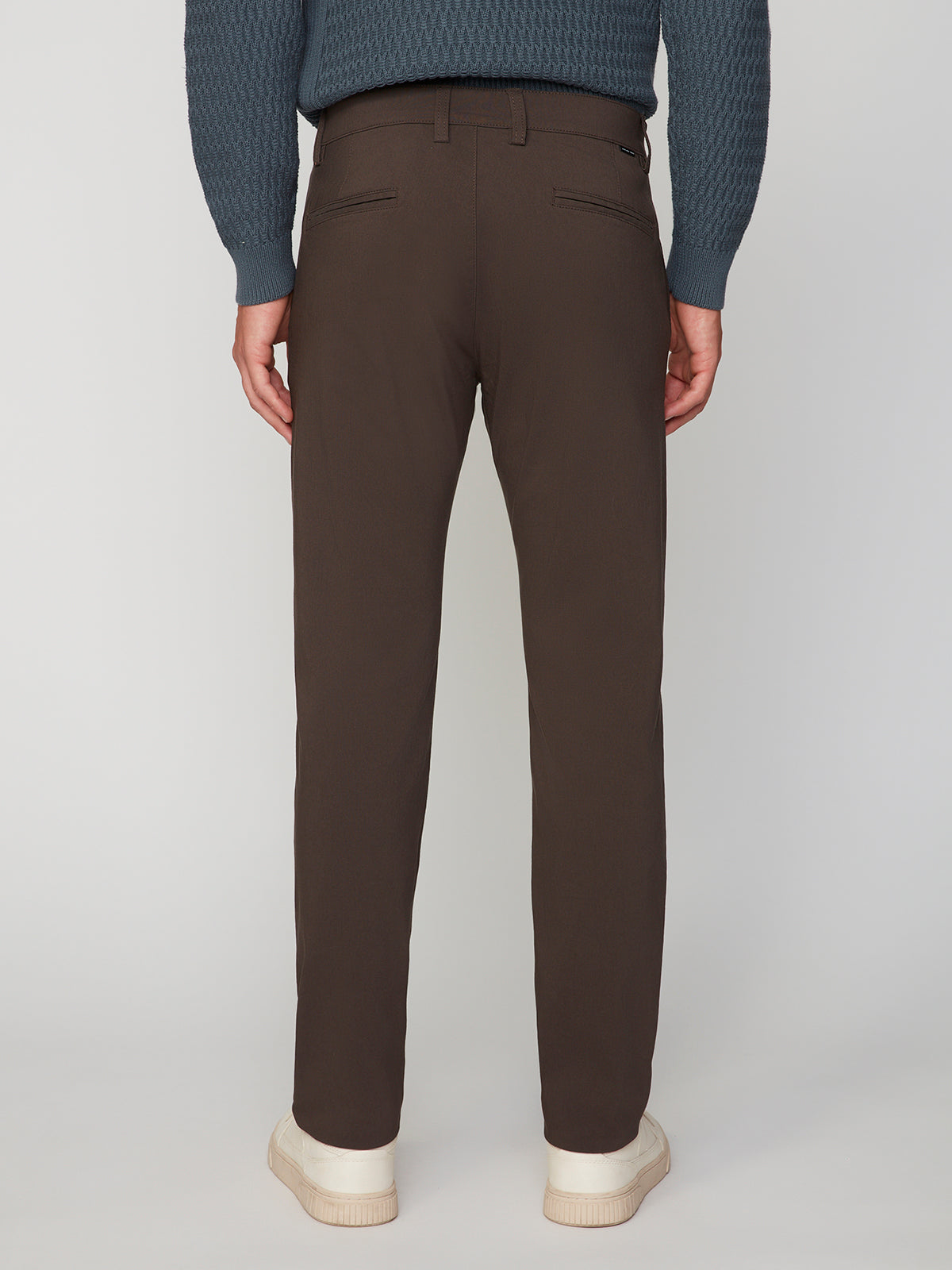 A person stands facing away, wearing PROJEK RAW MENS 5-POCKET SOLID BUNGEE CHINO PANT in dark brown, a textured blue sweater, and white sneakers against a plain light background.