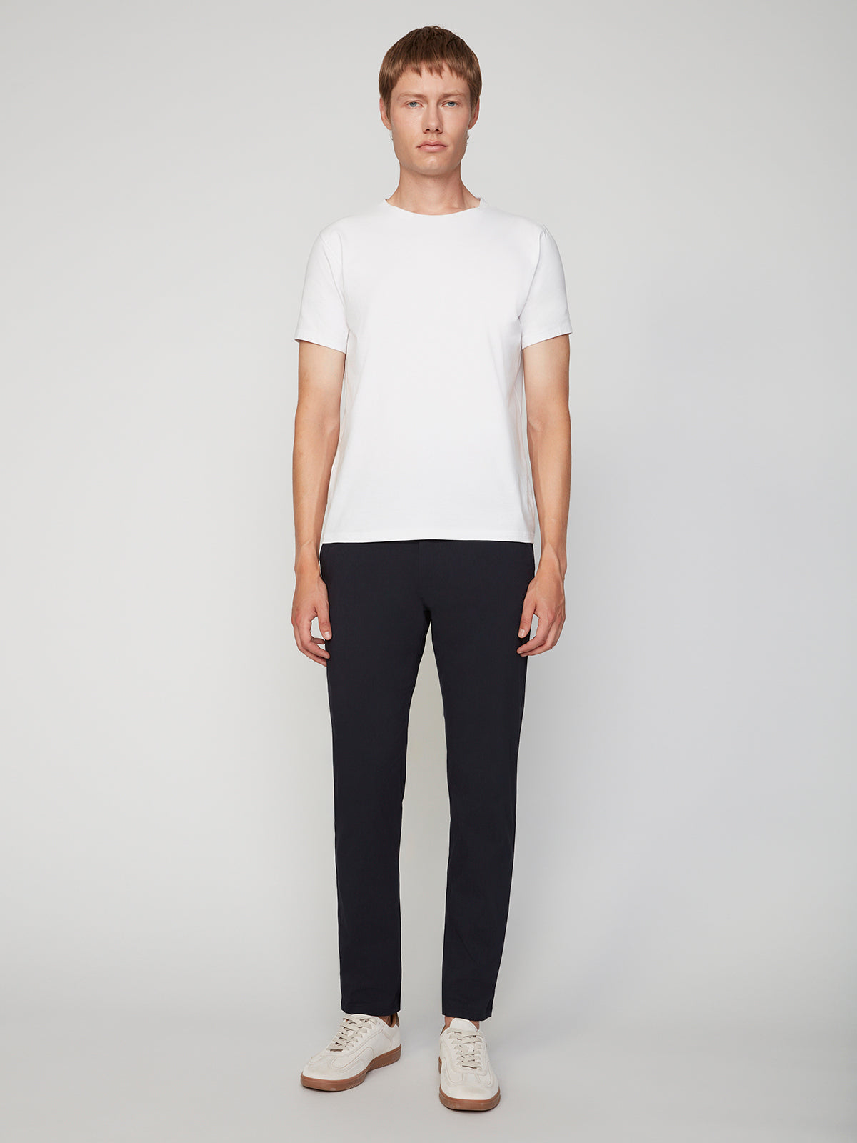 A person stands facing forward against a plain background, wearing a white short-sleeve t-shirt, PROJEK RAW MENS 5-POCKET SOLID BUNGEE CHINO PANT, and white sneakers.