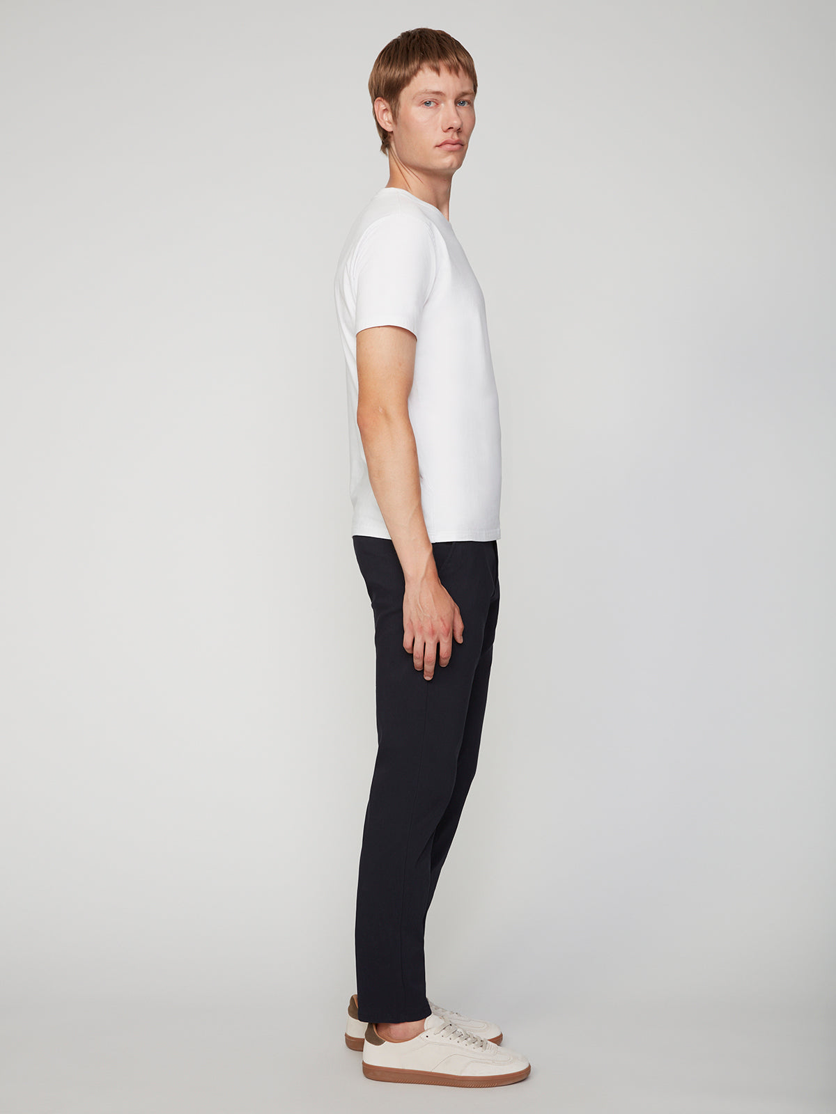 A man in profile stands against a plain light background, wearing a white tee, PROJEK RAW MENS 5-POCKET SOLID BUNGEE CHINO PANT, and white sneakers. He looks ahead neutrally with arms relaxed at his sides.