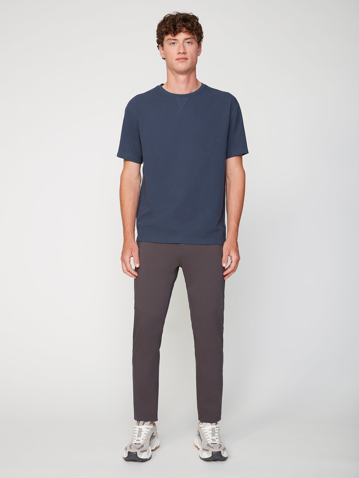A young man with short curly hair stands against a white background, wearing a navy blue short-sleeve t-shirt, PROJEK RAW MENS SOLID BUNGEE FASHION CHINO PANT in a tailored fit, and white athletic sneakers.