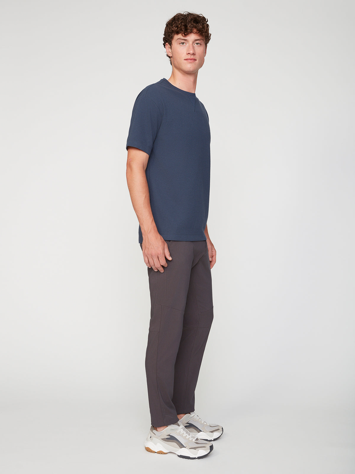 A young man with short curly brown hair wears a navy short-sleeve shirt, PROJEK RAW MENS SOLID BUNGEE FASHION CHINO PANT in a tailored fit, and white sneakers, standing sideways and looking at the camera against a white background.