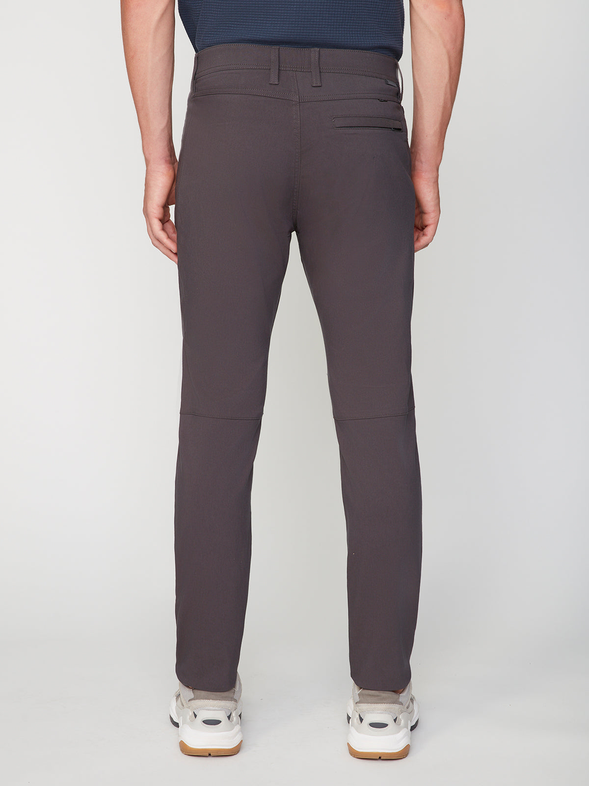 A man stands facing away, wearing PROJEK RAW MENS SOLID BUNGEE FASHION CHINO PANT in dark gray with white sneakers, his arms relaxed at his sides against a plain white background.