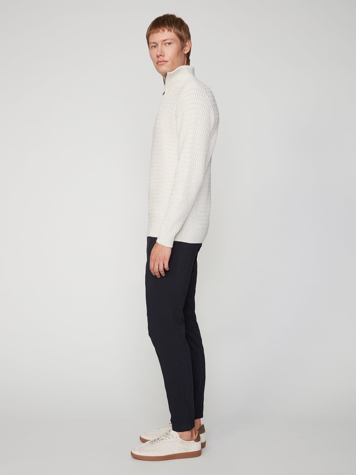 A person stands in profile against a plain background, wearing a white textured zip-neck sweater, PROJEK RAW MENS SOLID BUNGEE JOGGER pants with an elasticized waistband, and white sneakers with brown soles.