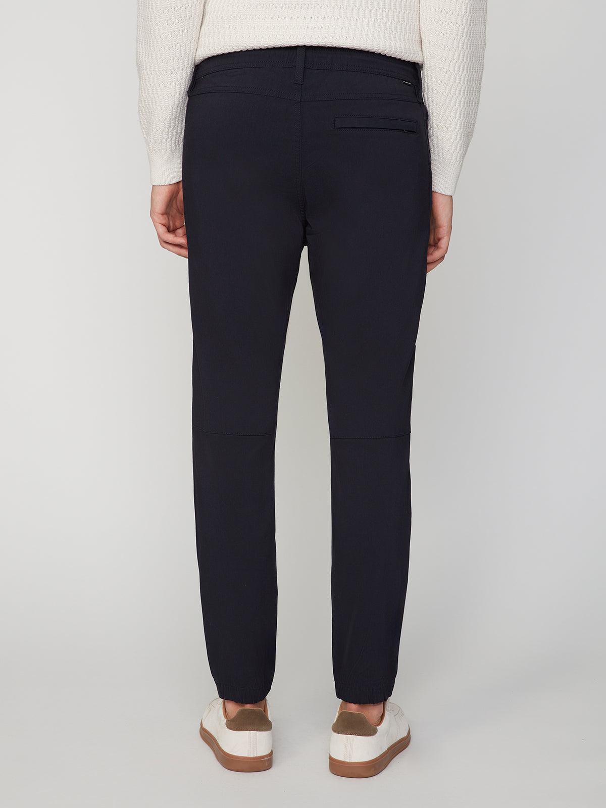 A person is shown from the back against a plain light background wearing PROJEK RAW MENS SOLID BUNGEE JOGGER pants in dark navy, paired with a cream sweater and white sneakers with brown soles.