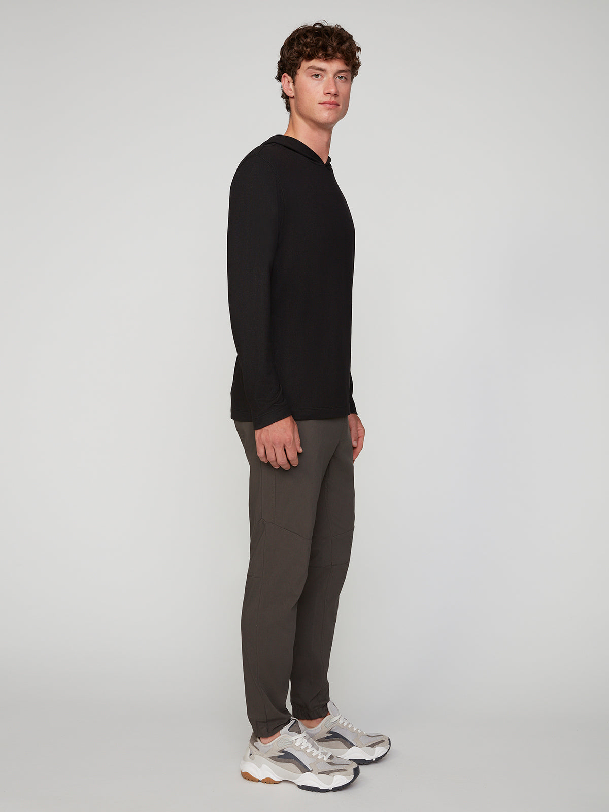 A young man stands sideways, facing the camera in a long-sleeve black shirt and PROJEK RAW MENS SOLID BUNGEE JOGGER pants with an elastic waist and modern fit, paired with white chunky-soled sneakers against a light gray background.