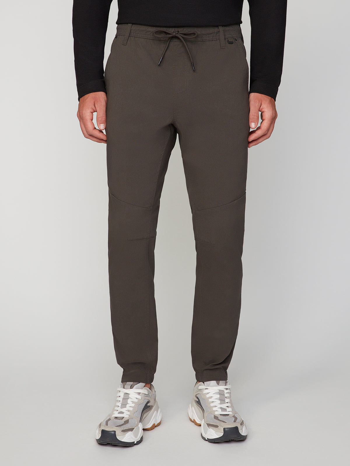 A person is wearing PROJEK RAW MENS SOLID BUNGEE JOGGER pants with an elasticized waistband, paired with a black long-sleeve shirt and white-gray sneakers, standing against a plain light background.