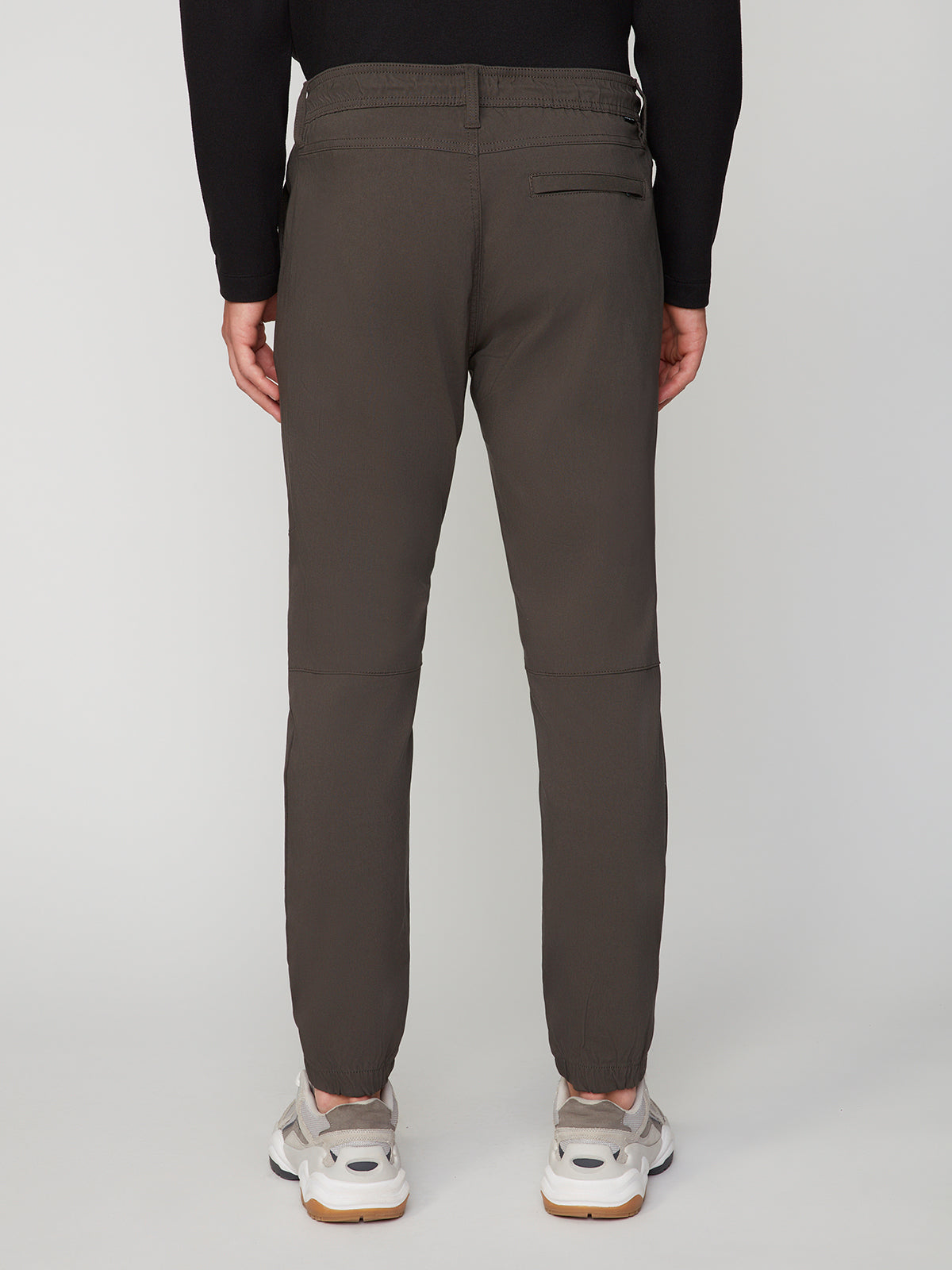 A person is seen from the back wearing PROJEK RAW MENS SOLID BUNGEE JOGGER pants with a modern fit and elasticized waistband, paired with white sneakers, standing against a plain light gray background.