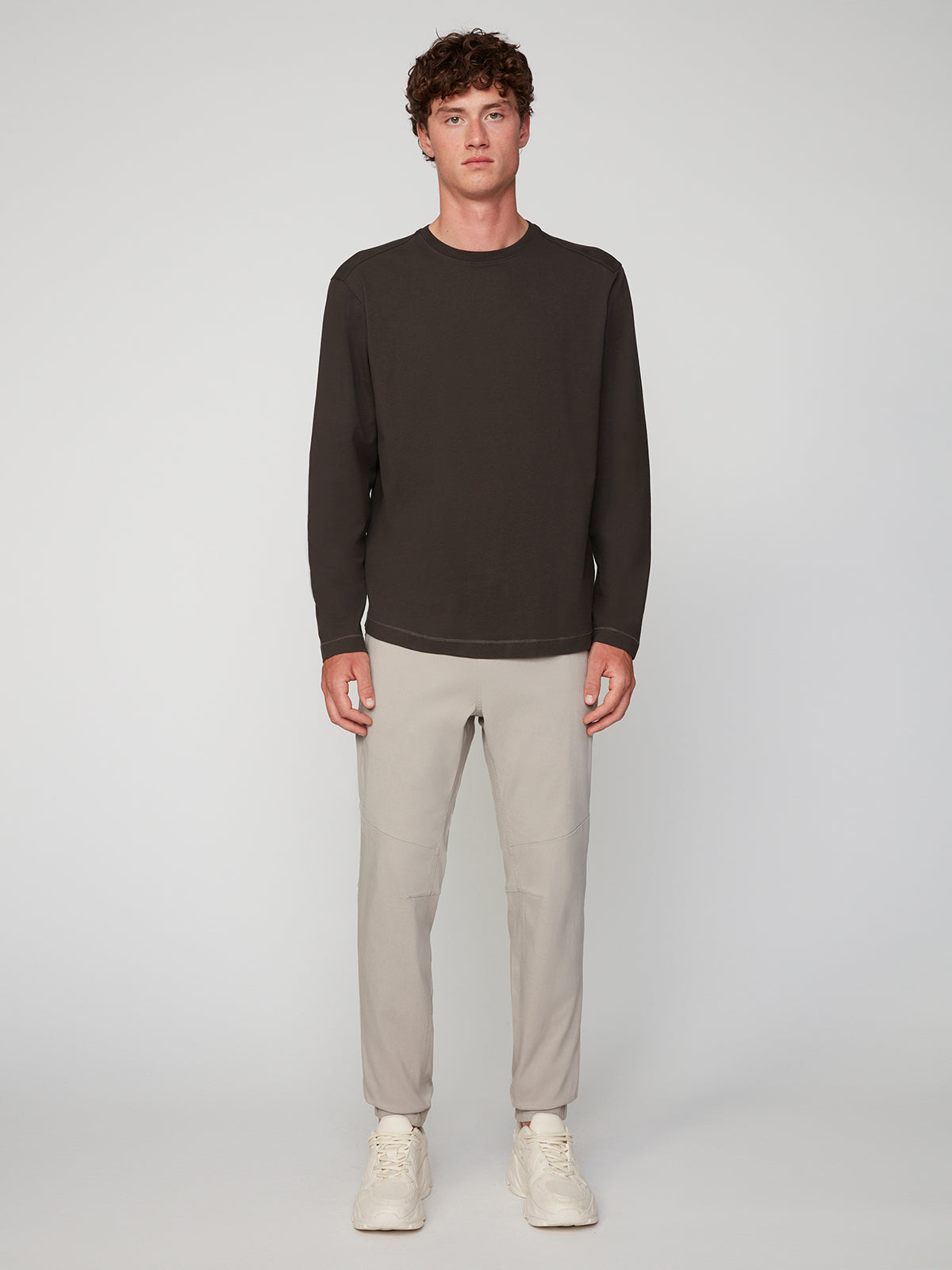 A young man with curly brown hair stands facing forward, wearing a long-sleeve dark brown shirt, PROJEK RAW MENS SOLID BUNGEE JOGGER in light beige, and off-white sneakers against a plain light background.