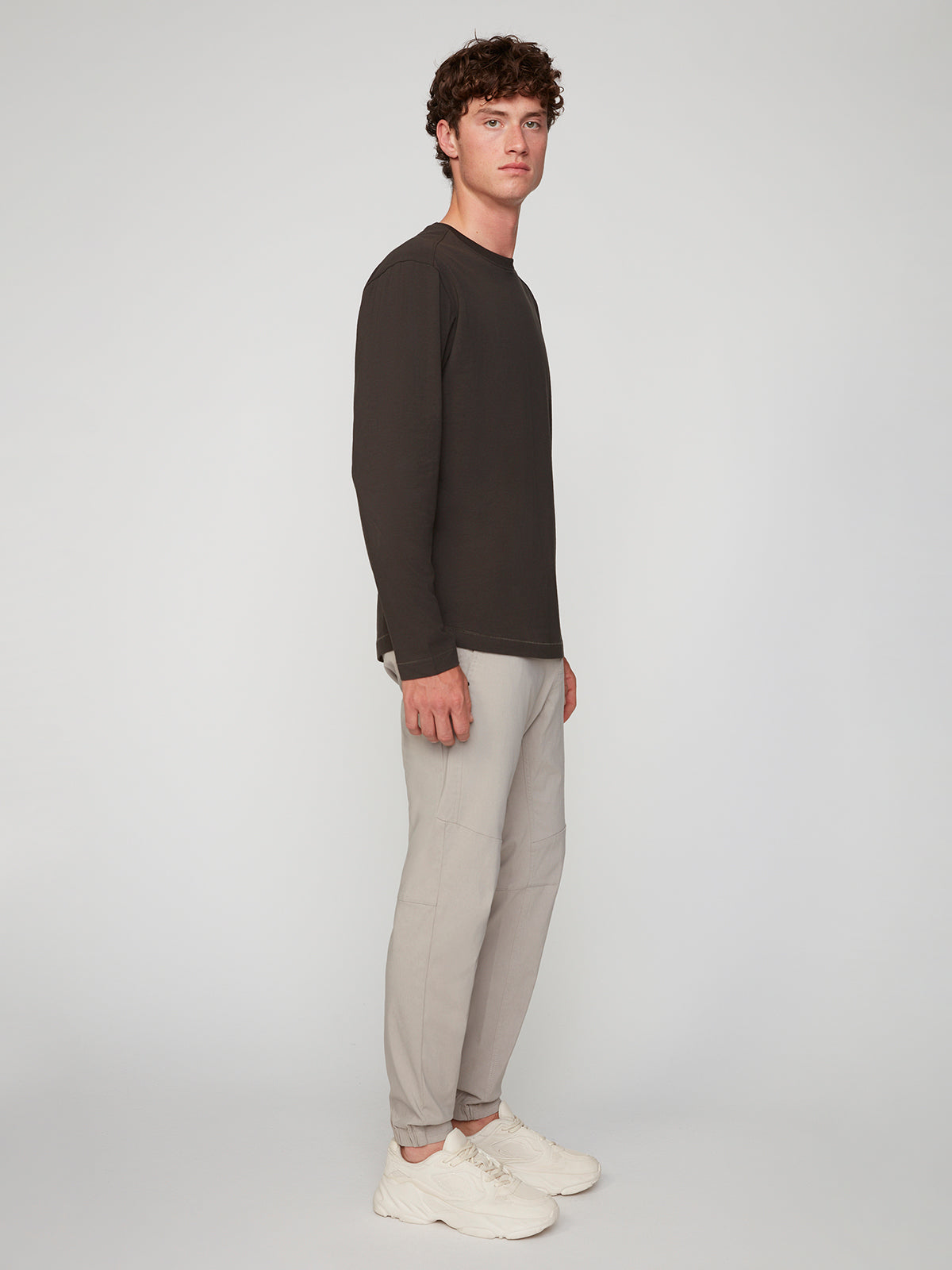 A young man with short curly hair stands sideways, sporting a dark long-sleeve shirt, PROJEK RAW MENS SOLID BUNGEE JOGGER with an elastic waistband, and white sneakers against a plain light background.