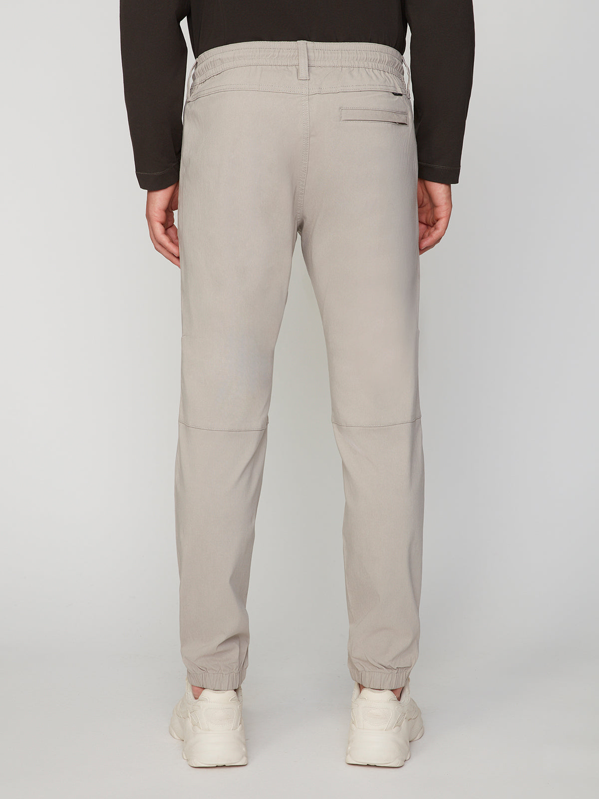 A person faces away, wearing PROJEK RAW MENS SOLID BUNGEE JOGGER pants in light gray with an elastic waistband and cuffs, paired with a black long-sleeve top and white sneakers against a plain light background.