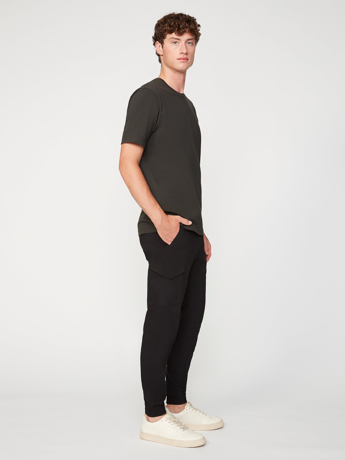 A young man wearing PROJEK RAW’s MENS SOLID BUNGEE CARGO JOGGER stands sideways with hands in his cargo pockets, paired with a dark tee and white sneakers against a simple light background.