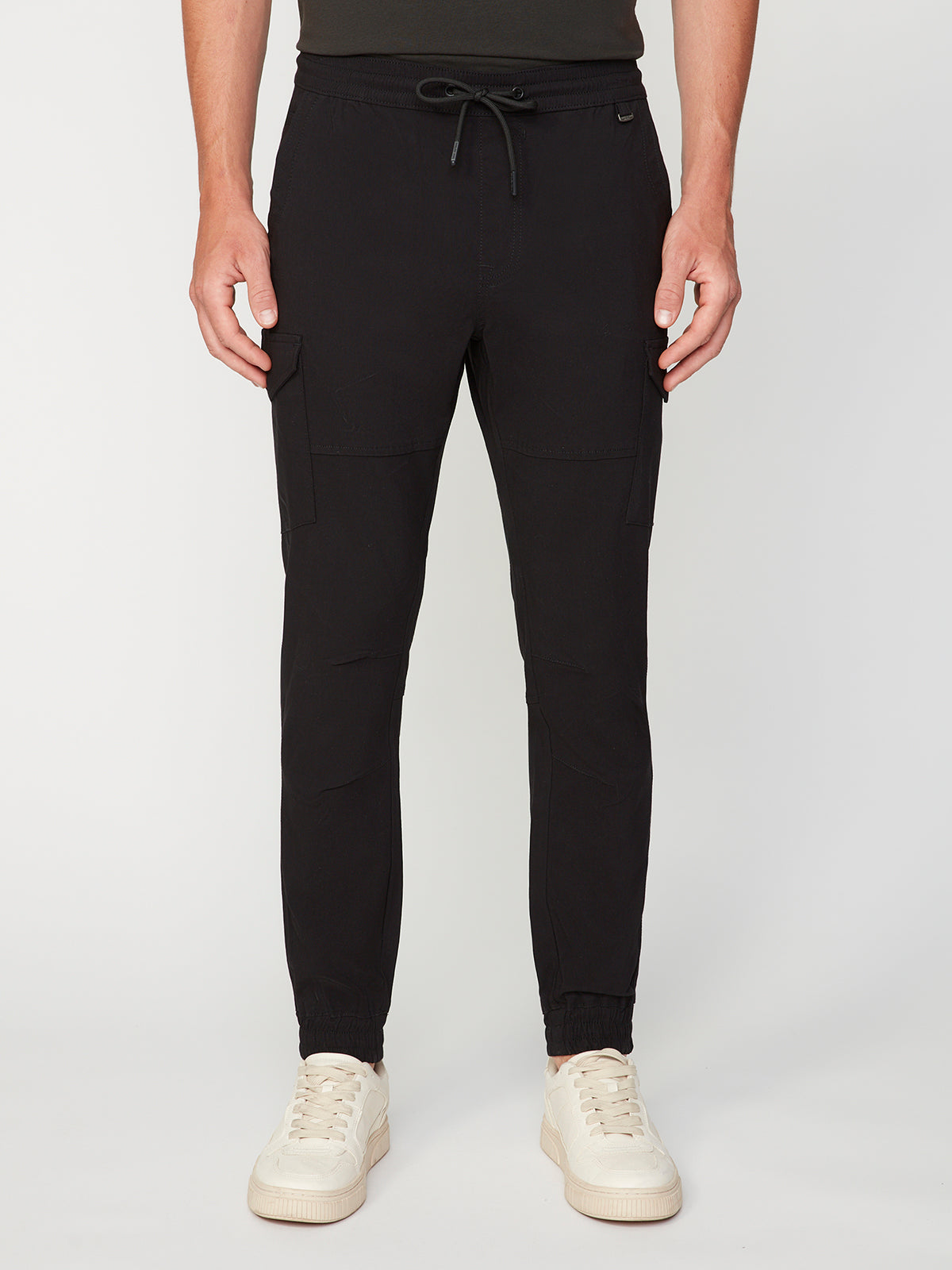 A person wears PROJEK RAW MENS SOLID BUNGEE CARGO JOGGER pants with cargo pockets and an elasticized waistband, paired with cream sneakers, standing against a plain white background.