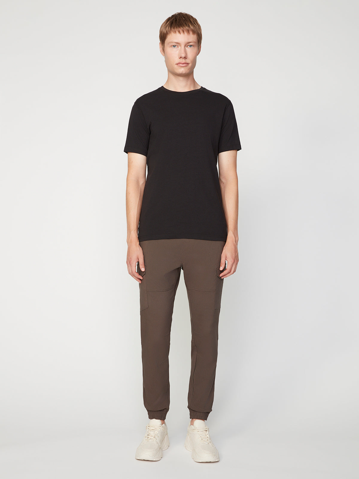 A person faces forward against a plain background, wearing PROJEK RAW MENS SOLID BUNGEE CARGO JOGGER pants in brown with an elasticized waistband and cargo pockets, paired with a black short-sleeve t-shirt and white sneakers.