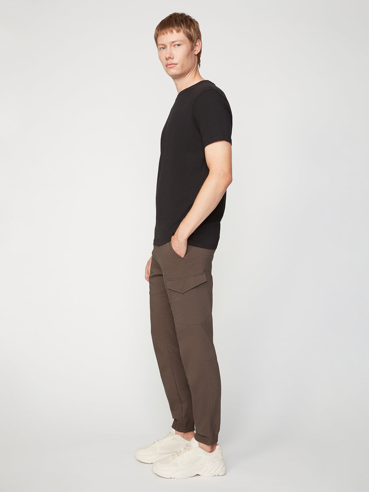 A person stands sideways against a plain light background, wearing the PROJEK RAW MENS SOLID BUNGEE CARGO JOGGER with cargo pockets and elasticized waistband, a black short-sleeve shirt, and white sneakers, with one hand in their pocket.