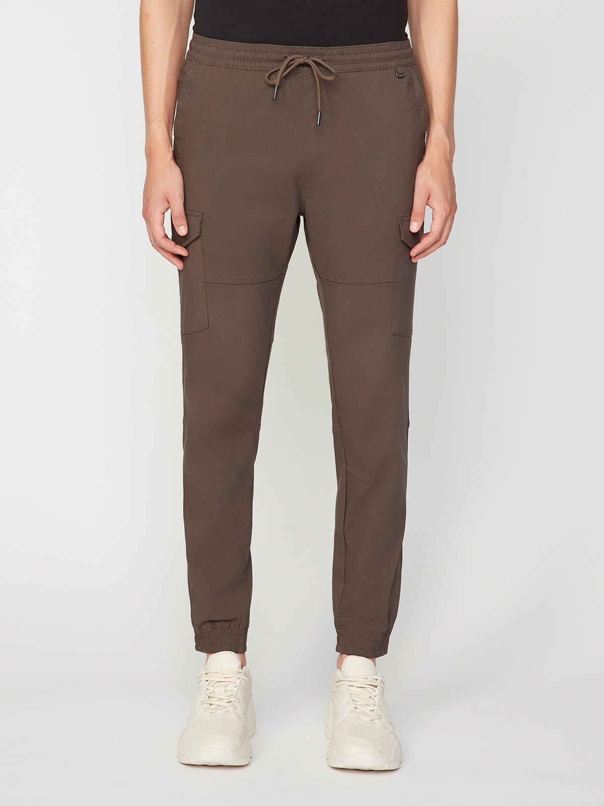 The lower half of a person wearing PROJEK RAW MENS SOLID BUNGEE CARGO JOGGER pants with cargo pockets and an elasticized waistband, paired with white sneakers, is shown against a plain white background.