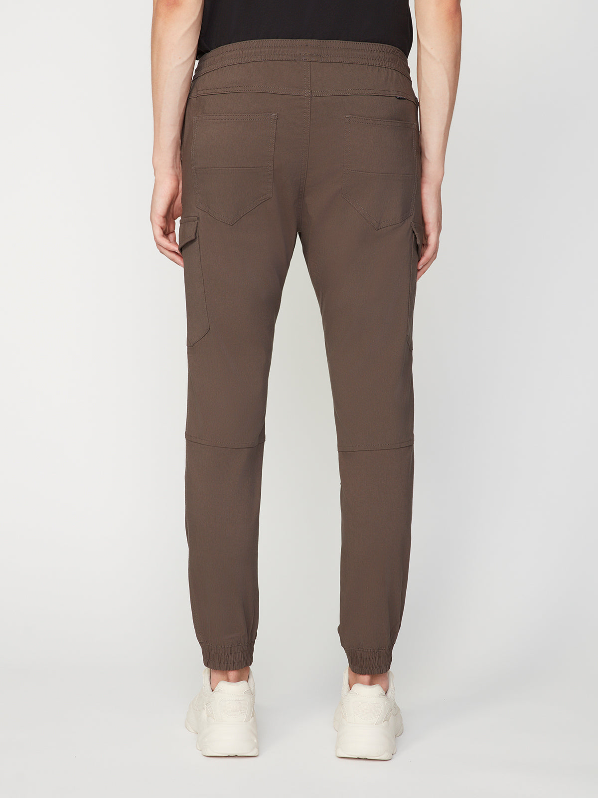 A person seen from the back wears PROJEK RAWs MENS SOLID BUNGEE CARGO JOGGER in brown, featuring cargo pockets and elastic ankle cuffs, paired with white sneakers. The background is plain white.
