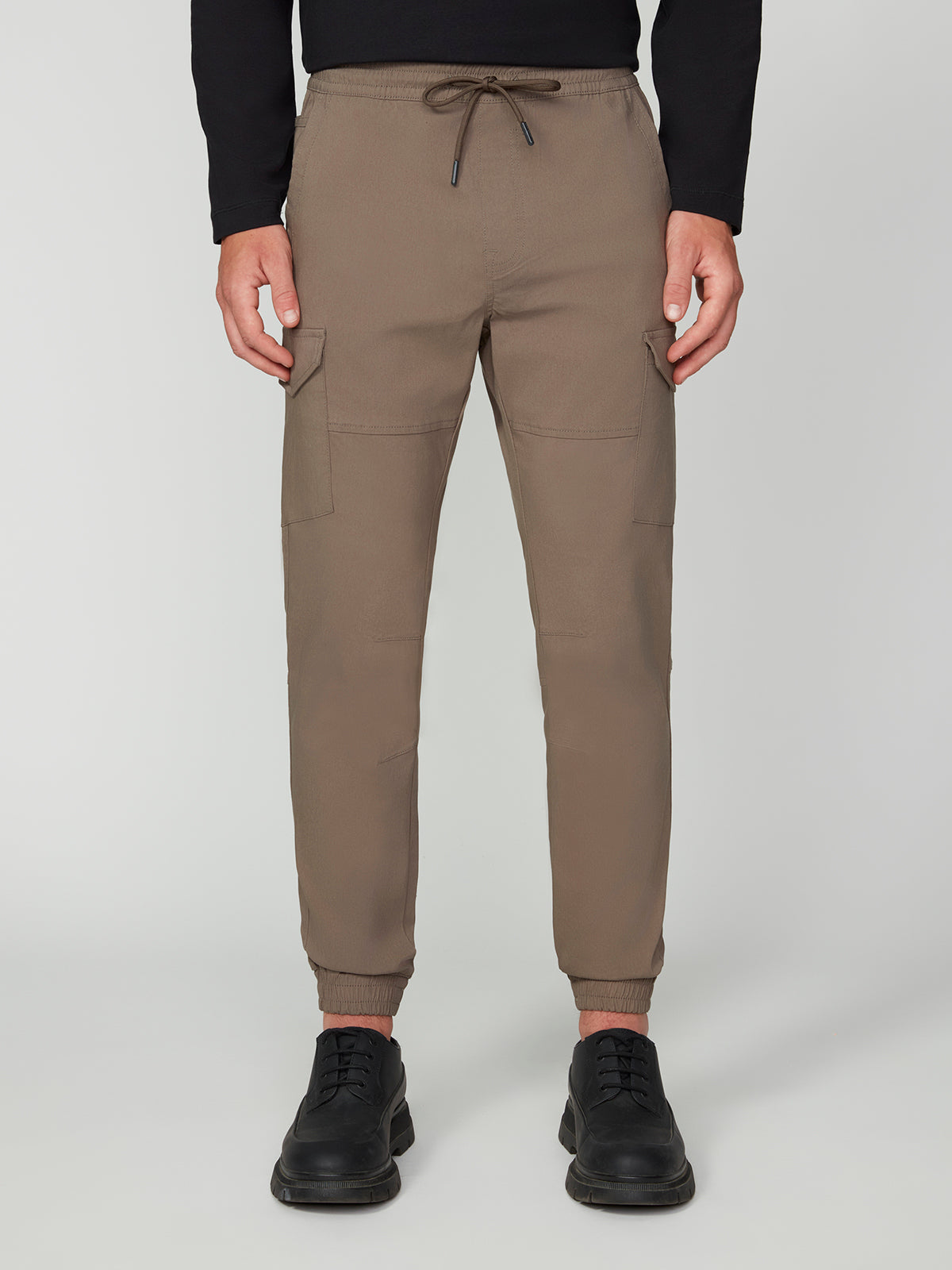 Wearing PROJEK RAW MENS SOLID BUNGEE CARGO JOGGER pants with thigh pockets and elastic cuffs, a black long-sleeve top, and black shoes, a person stands against a plain light gray background.