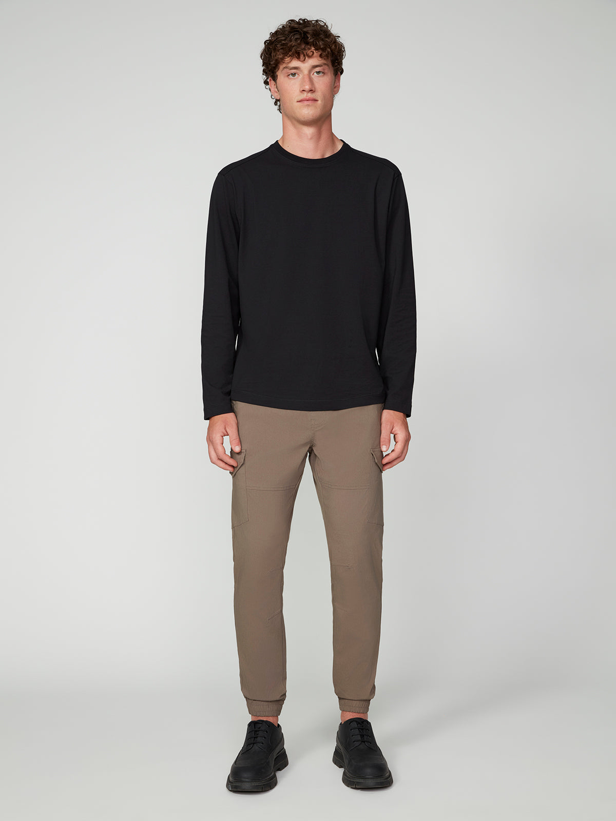 A young man with curly hair faces forward against a light gray background, wearing black shoes, a black long-sleeve shirt, and PROJEK RAW MENS SOLID BUNGEE CARGO JOGGER.
