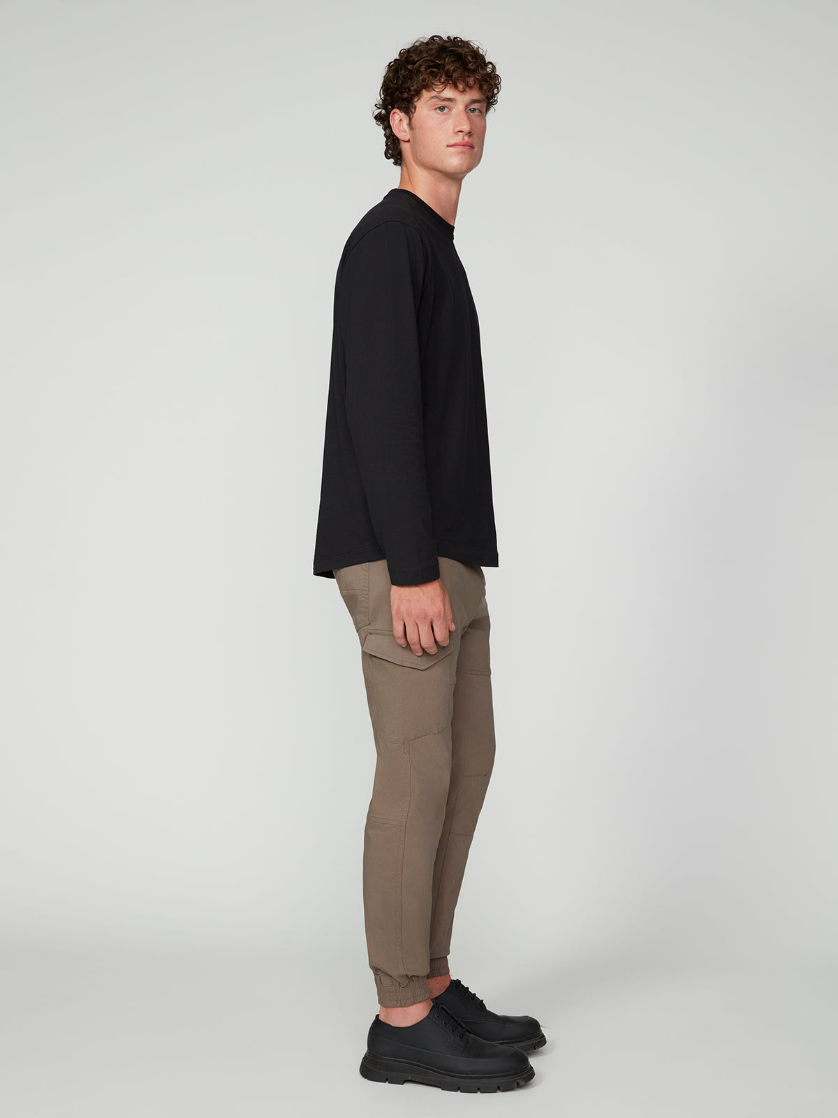 A young man with curly hair stands sideways in a black long-sleeve shirt, PROJEK RAW MENS SOLID BUNGEE CARGO JOGGER pants, and black shoes against a plain light gray background.