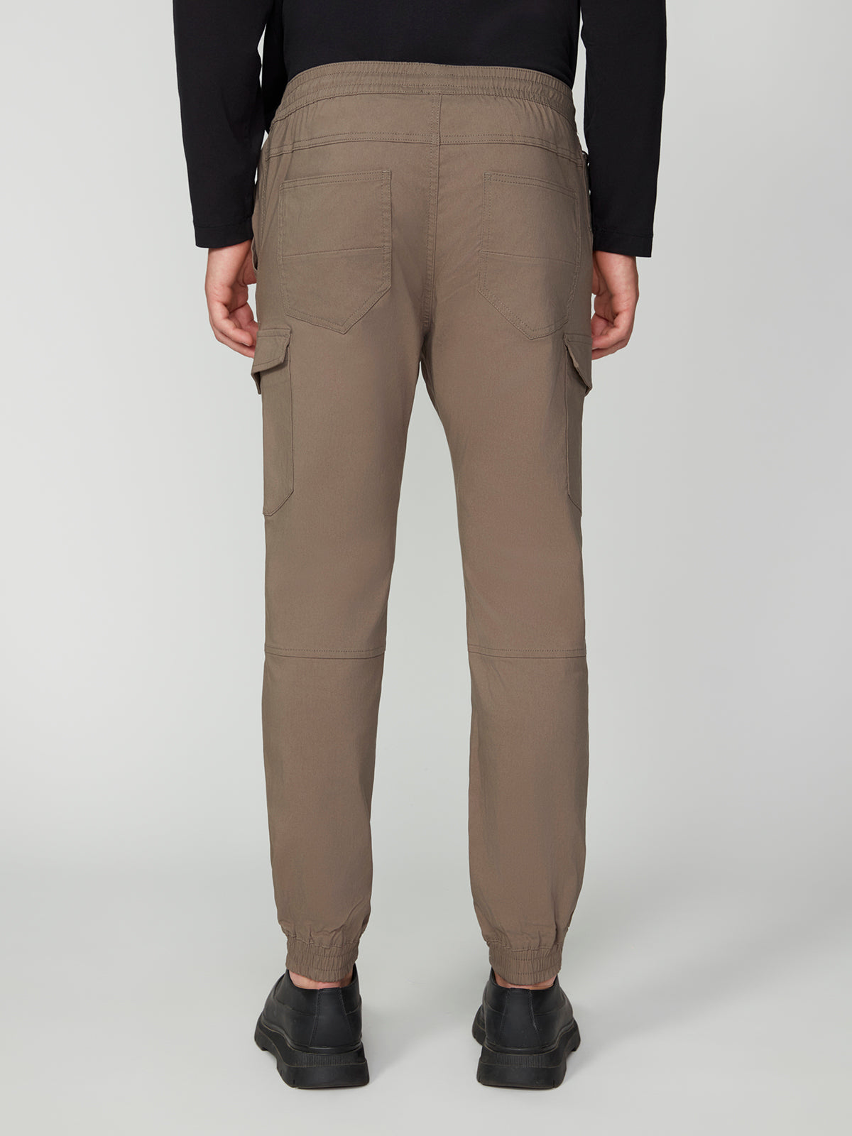 A person is shown from the back against a plain light background wearing PROJEK RAW MENS SOLID BUNGEE CARGO JOGGER pants in taupe with cargo pockets and elastic cuffs, paired with black shoes and a black long-sleeve shirt.