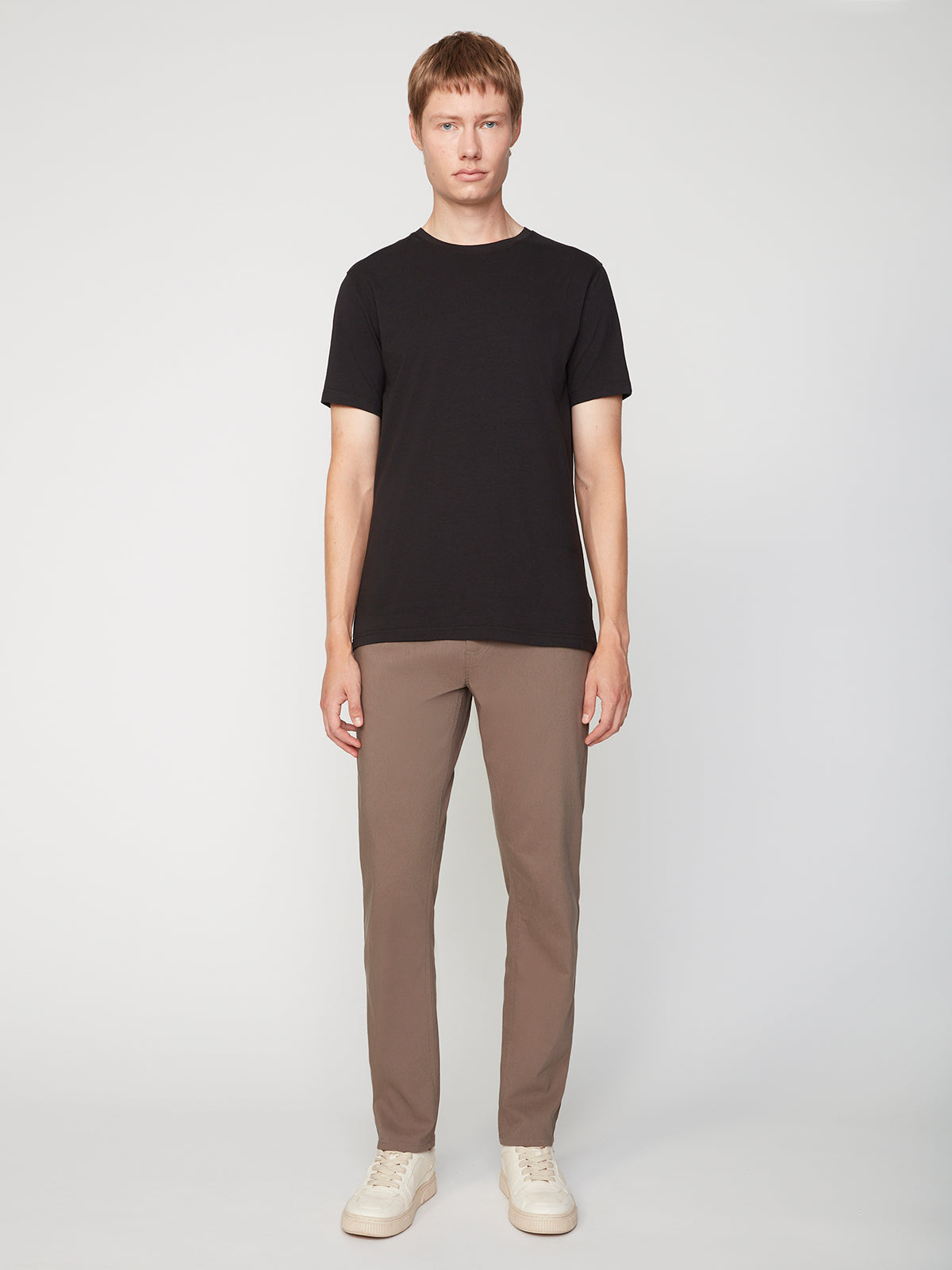 A young man with short light brown hair faces forward, wearing a plain black t-shirt, PROJEK RAW MENS SOLID BUNGEE RELAXED 5-POCKET PANT in light brown, and off-white sneakers against a plain light background.