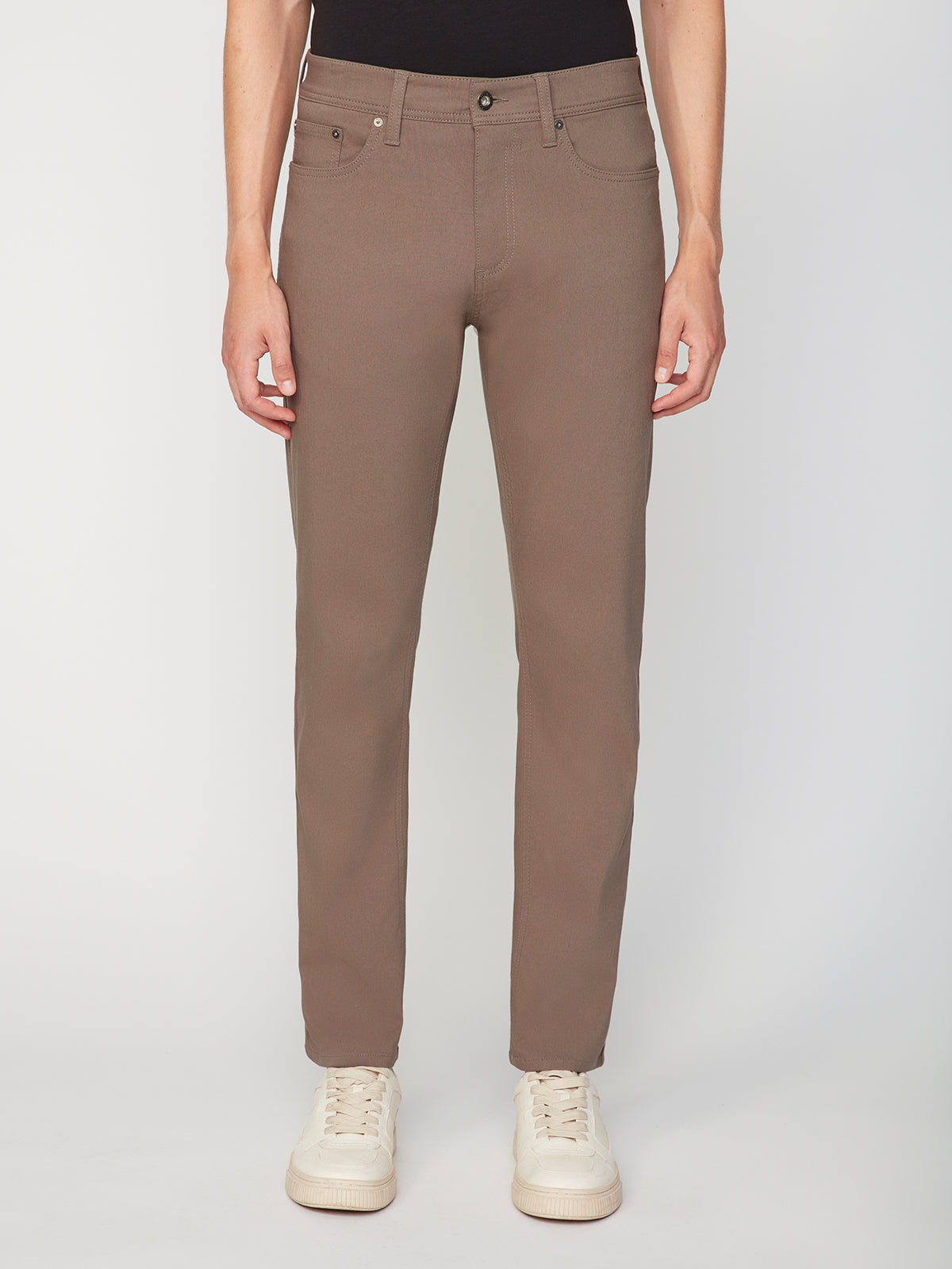 A person models the PROJEK RAW MENS SOLID BUNGEE RELAXED 5-POCKET PANT in light brown with white sneakers, lower torso and legs visible, standing against a plain white background.