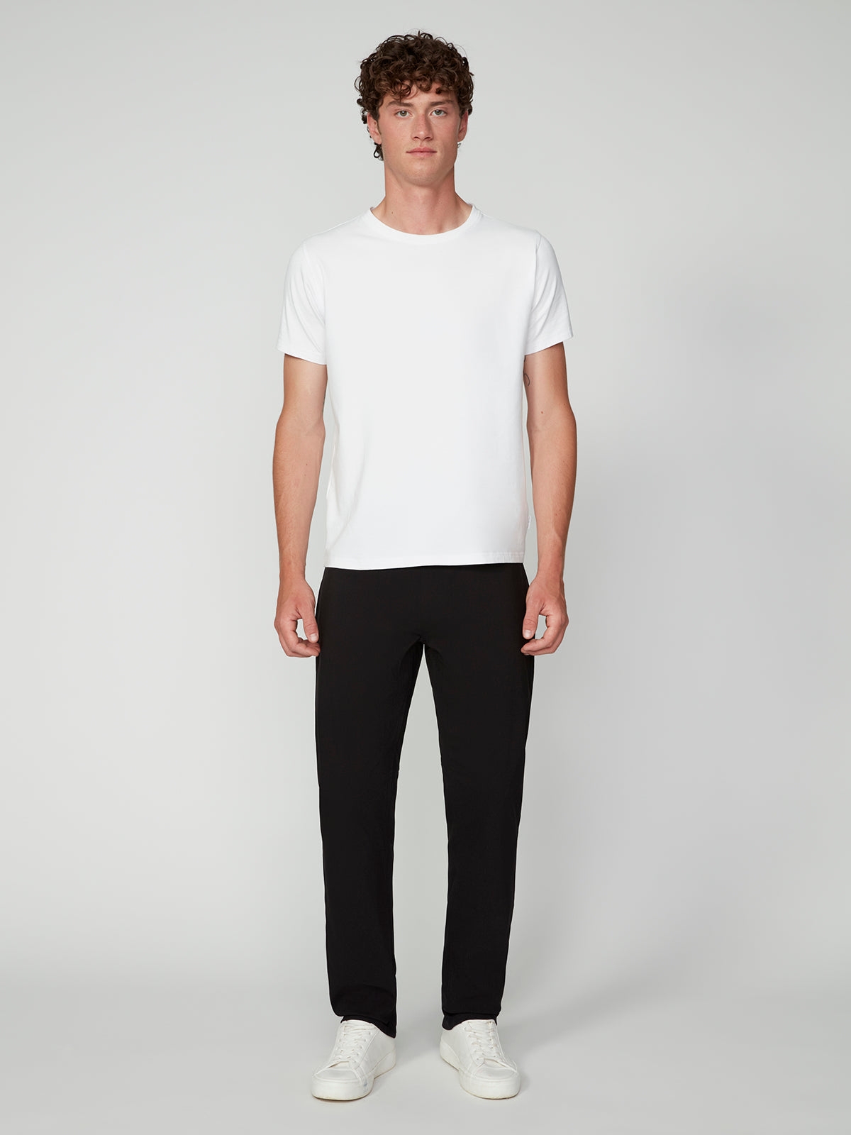 A young man with curly brown hair stands facing forward in a plain white T-shirt, PROJEK RAW MENS SOLID BUNGEE RELAXED 5-POCKET PANT in black, and white sneakers against a plain light gray background.