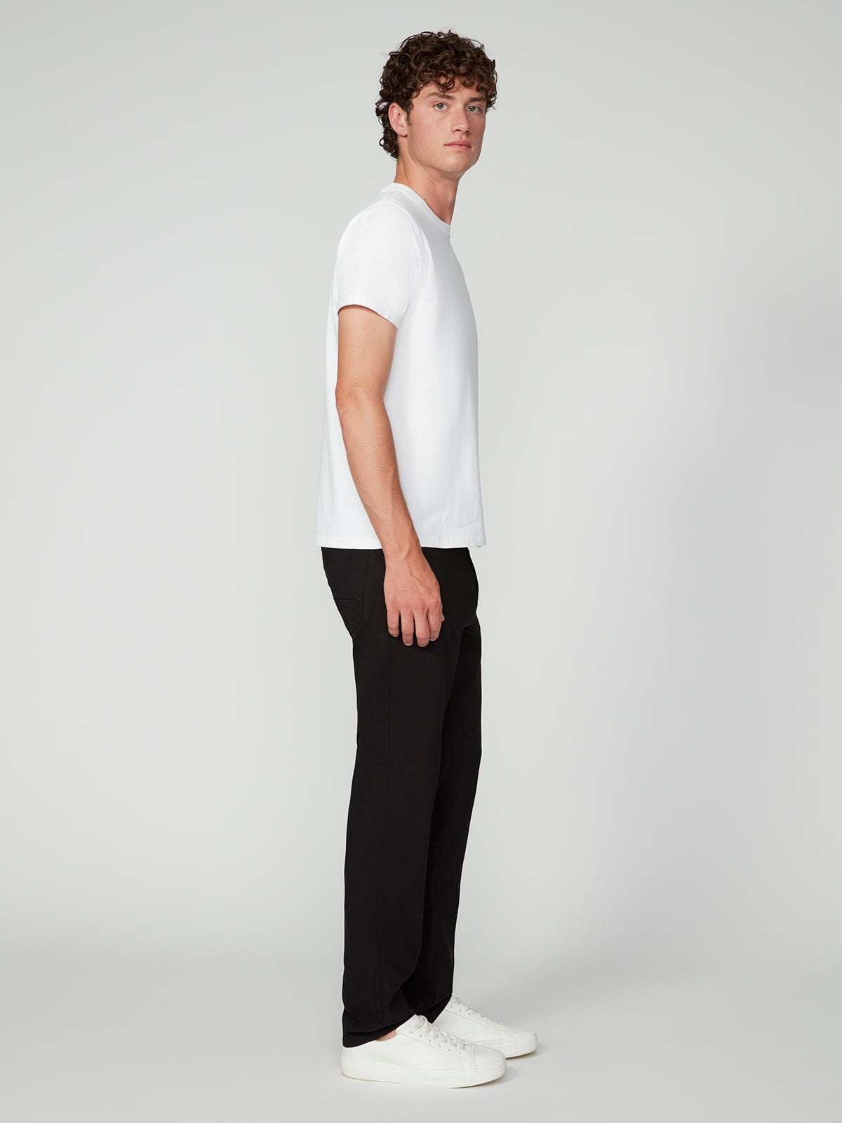 A young man with curly hair stands in profile against a plain light background, wearing a white t-shirt, PROJEK RAW MENS SOLID BUNGEE RELAXED 5-POCKET PANT with an elastic waistband, and white sneakers.