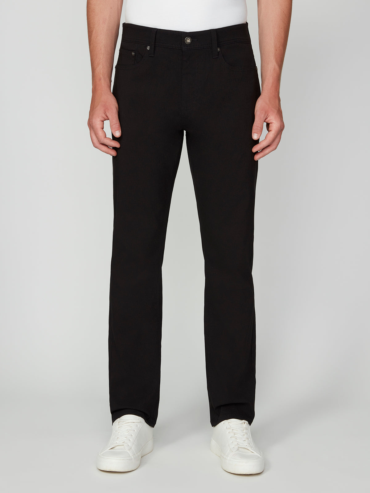 A man wears PROJEK RAW MENS SOLID BUNGEE RELAXED 5-POCKET PANT in black, paired with a white shirt and white sneakers, standing facing forward against a plain light background.