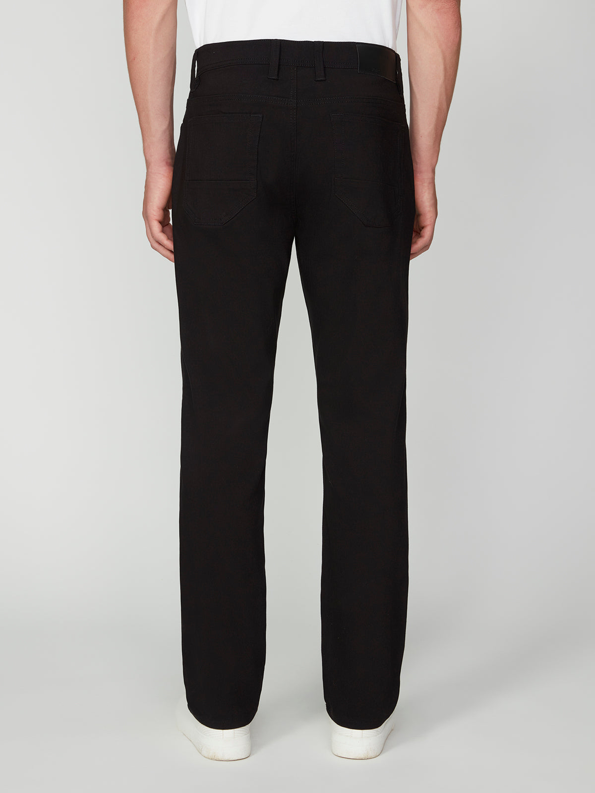 A person faces away from the camera, wearing PROJEK RAW MENS SOLID BUNGEE RELAXED 5-POCKET PANT in black, paired with a white shirt and white shoes, set against a plain light background.
