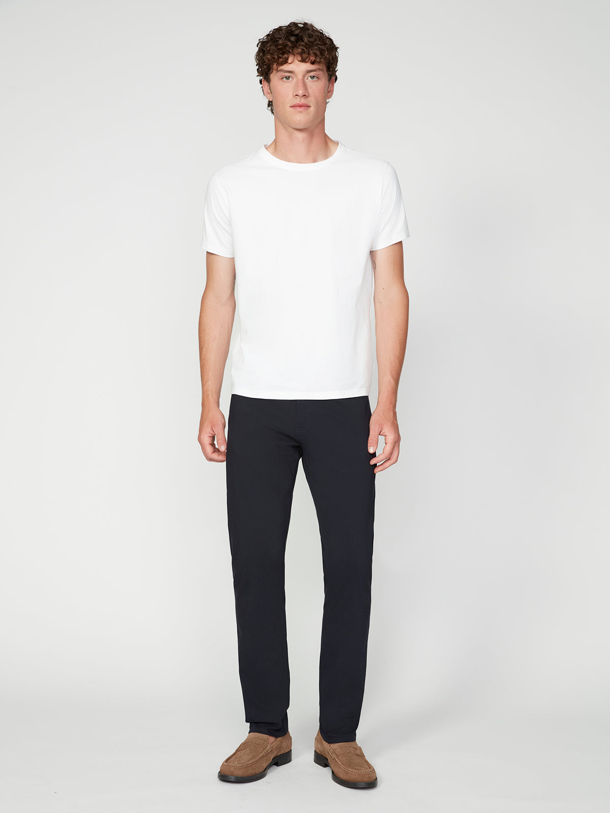 A young man with short curly hair faces forward, wearing a plain white t-shirt, PROJEK RAW MENS SOLID BUNGEE RELAXED 5-POCKET PANT, and brown loafers against a simple white background.