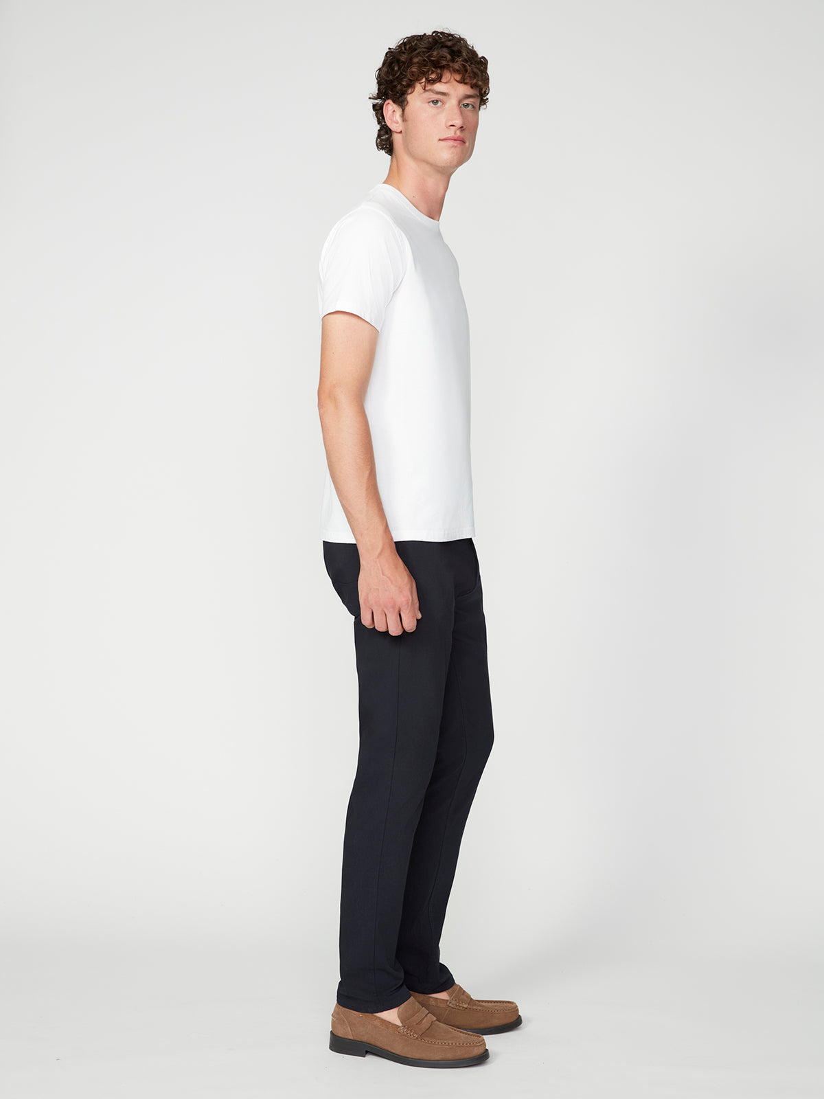 A man with curly hair stands sideways in a plain white t-shirt, brown loafers, and PROJEK RAW MENS SOLID BUNGEE RELAXED 5-POCKET PANT against a plain white background.