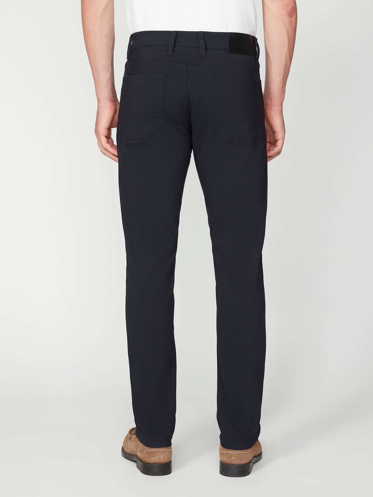 A man is shown from behind against a plain white background, wearing PROJEK RAW MENS SOLID BUNGEE RELAXED 5-POCKET PANT in black, a tucked-in white shirt, and brown shoes.