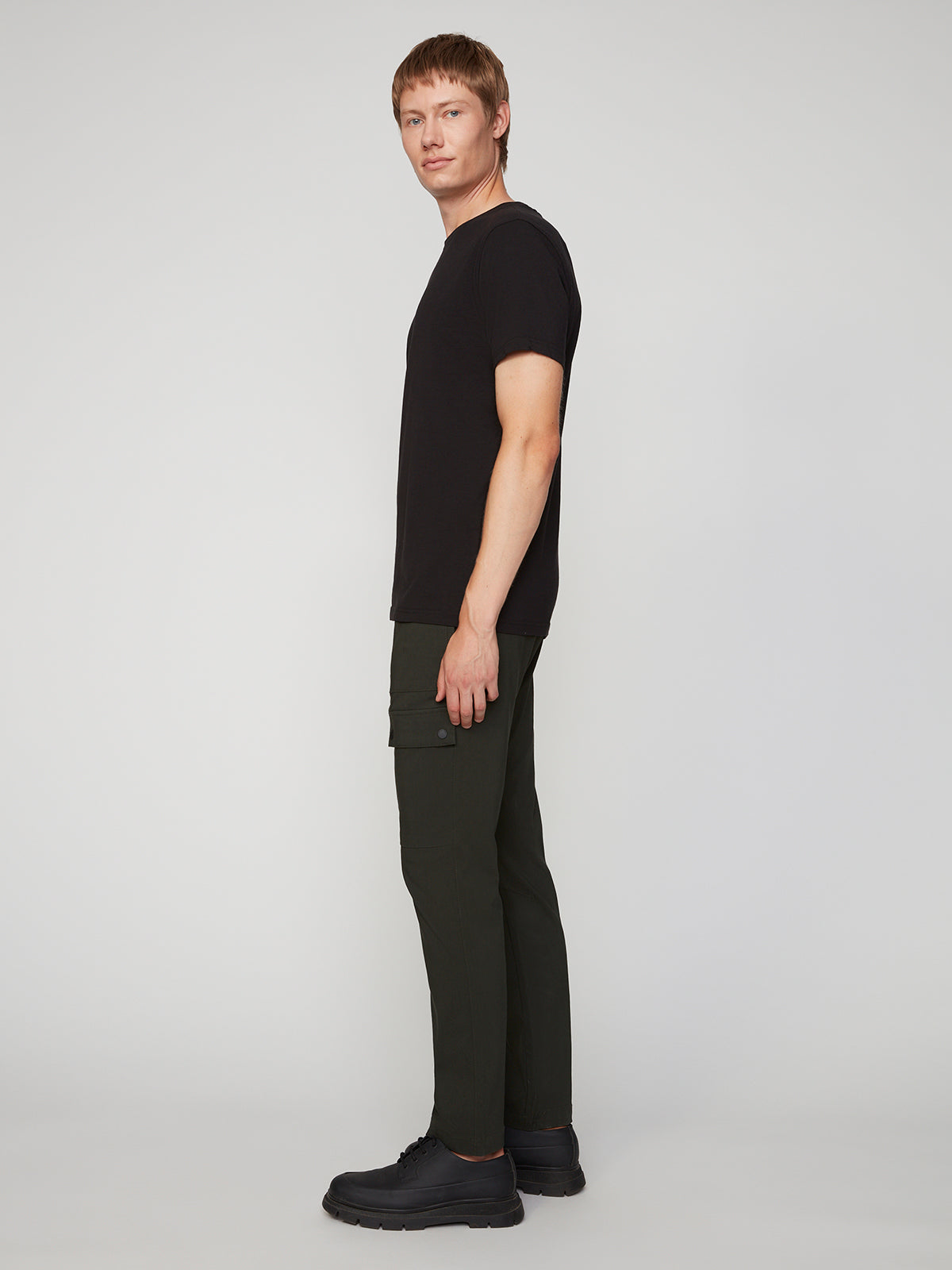 A person with short light brown hair stands in profile against a plain light background, wearing a black t-shirt, black shoes, and PROJEK RAW MENS SOLID BUNGEE RELAXED CARGO PANT.