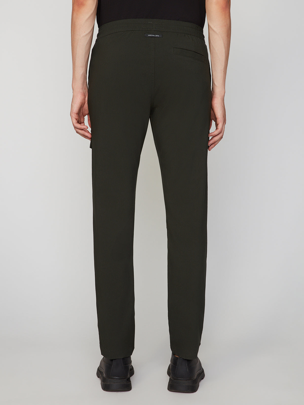 A person stands with their back to the camera, wearing black shoes, a black shirt, and PROJEK RAW MENS SOLID BUNGEE RELAXED CARGO PANT, set against a plain light background.