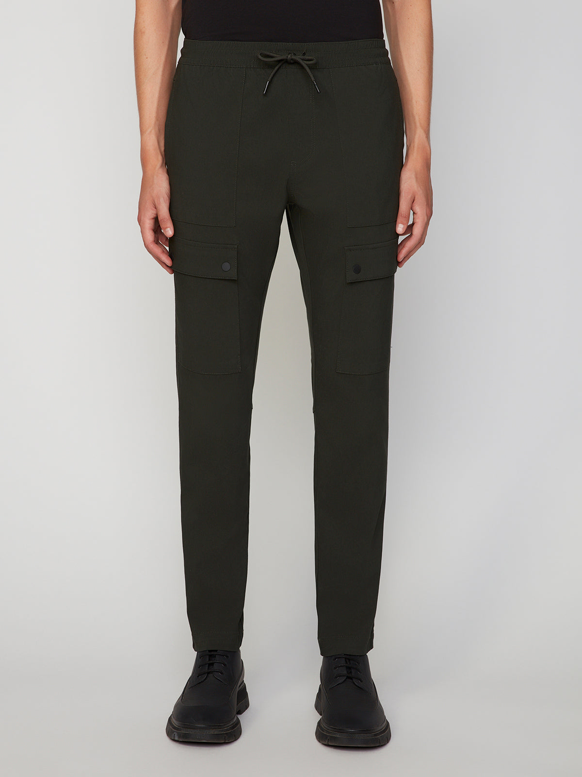 A person wears PROJEK RAW MENS SOLID BUNGEE RELAXED CARGO PANT in dark green, featuring a bungee waistband and buttoned flap thigh pockets, paired with black dress shoes against a plain light background.