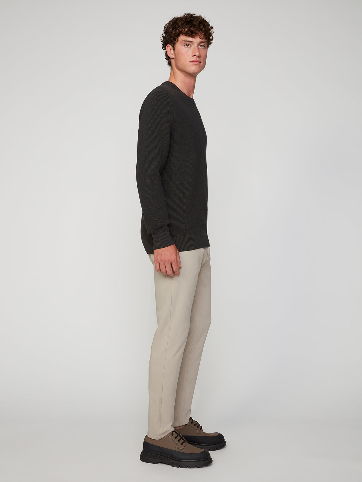 A young man with curly hair stands sideways, facing the camera. He wears PROJEK RAW’s black sweater and MEN’S RELAXED BUNGEE TWILL CHINO PANT, paired with brown lace-up shoes. The background is plain and light-colored.