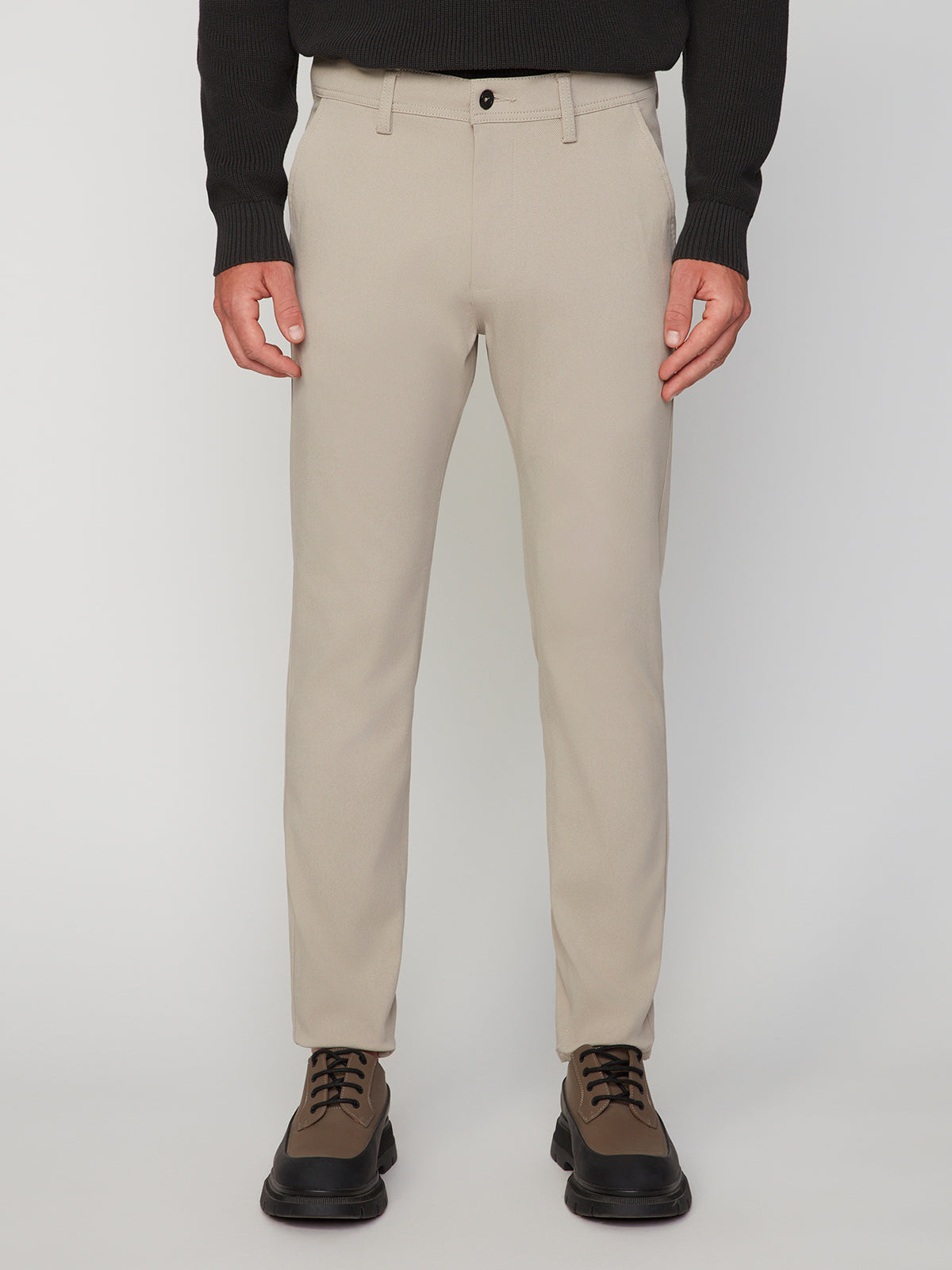 A person wearing PROJEK RAW MEN’S RELAXED BUNGEE TWILL CHINO PANT, a black long-sleeve shirt, and brown lace-up shoes stands against a plain white background. The photo is cropped from the shoulders down.