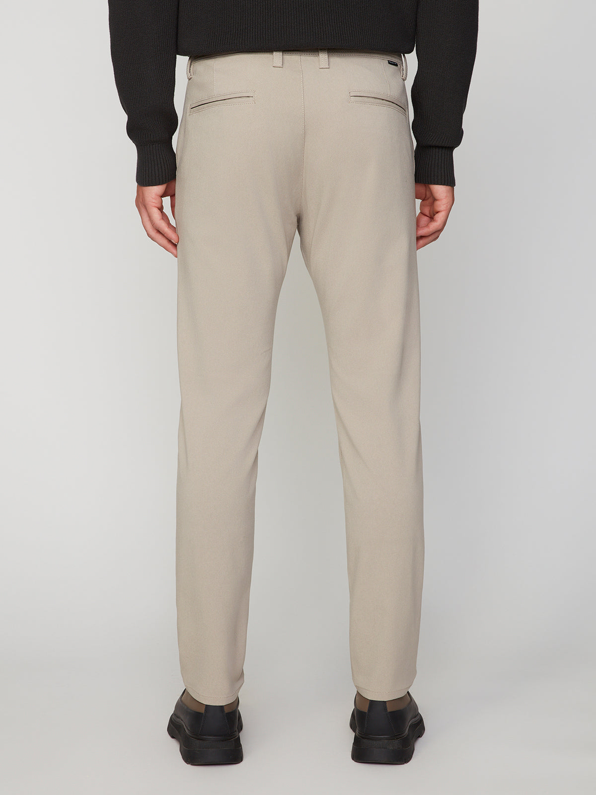 A person is shown from the back, standing against a plain white background, wearing PROJEK RAW MEN’S RELAXED BUNGEE TWILL CHINO PANT in beige, paired with a black sweater and black shoes.