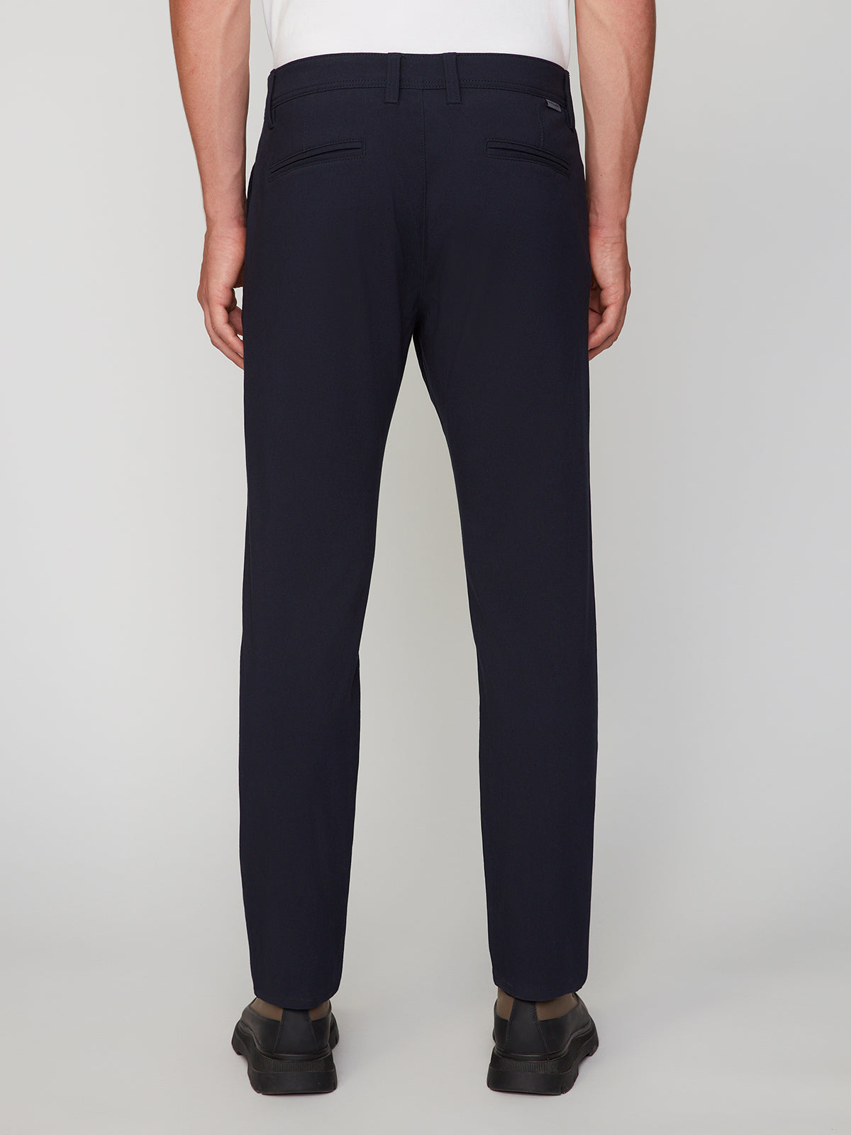 A person wearing PROJEK RAW MEN’S RELAXED BUNGEE TWILL CHINO PANT in dark navy with black shoes is seen from the back against a plain light gray background.