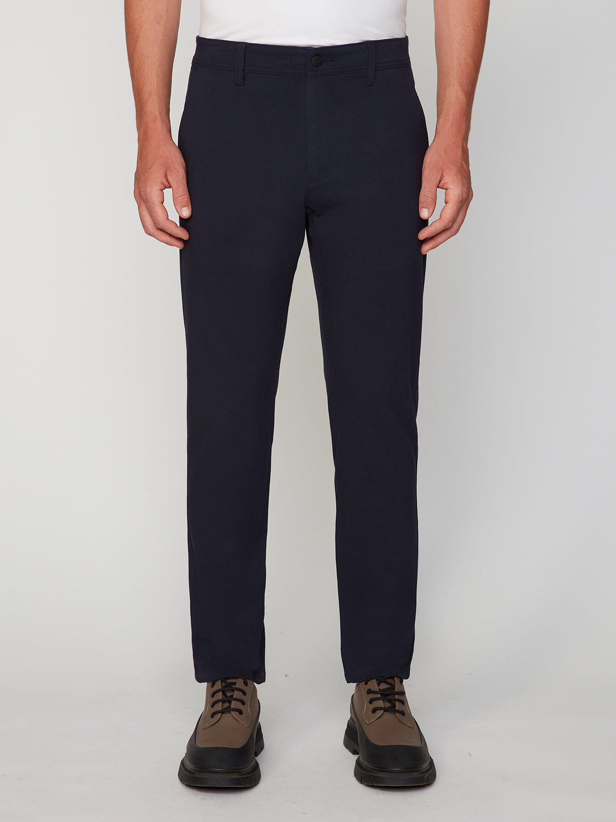 A person wears PROJEK RAW MEN’S RELAXED BUNGEE TWILL CHINO PANT in dark navy, paired with brown lace-up shoes. The photo, cropped from the waist down, features the person standing against a plain white background.